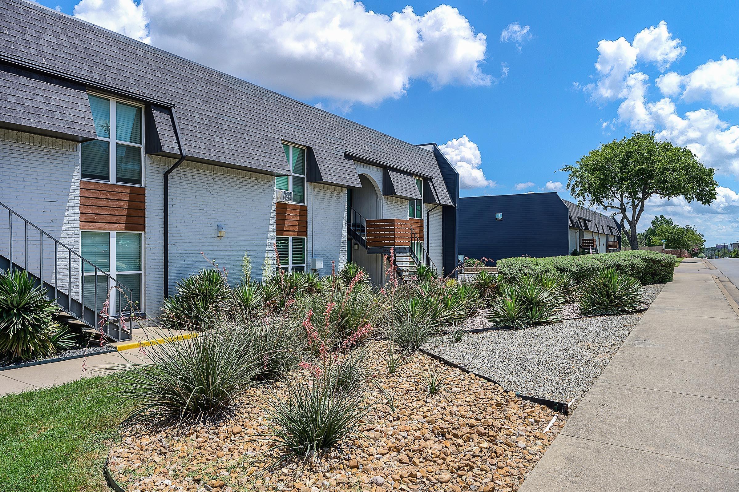 A landscaped apartment complex featuring two-story buildings with a mix of white and dark exteriors. The foreground includes low shrubs and ornamental grasses, while a concrete walkway runs alongside. The sky is partly cloudy, adding to the bright and inviting atmosphere of the setting.