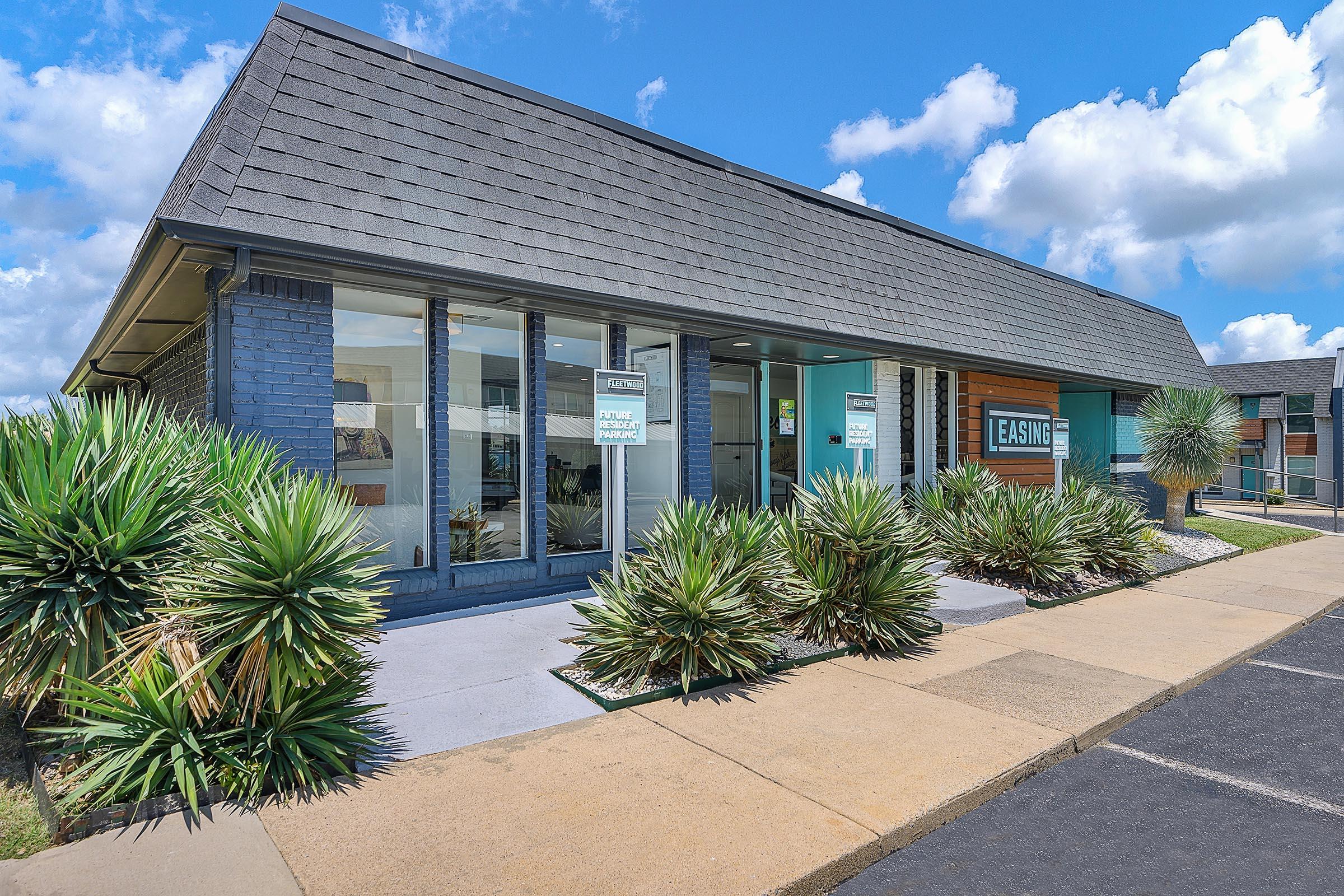 A modern single-story building with a gray shingle roof and large windows. The facade features a mix of blue and gray colors, with a sign that reads "EASING." Surrounding the building are neatly trimmed palm-like plants and a paved path leading to the entrance. The sky is partly cloudy with blue hues.