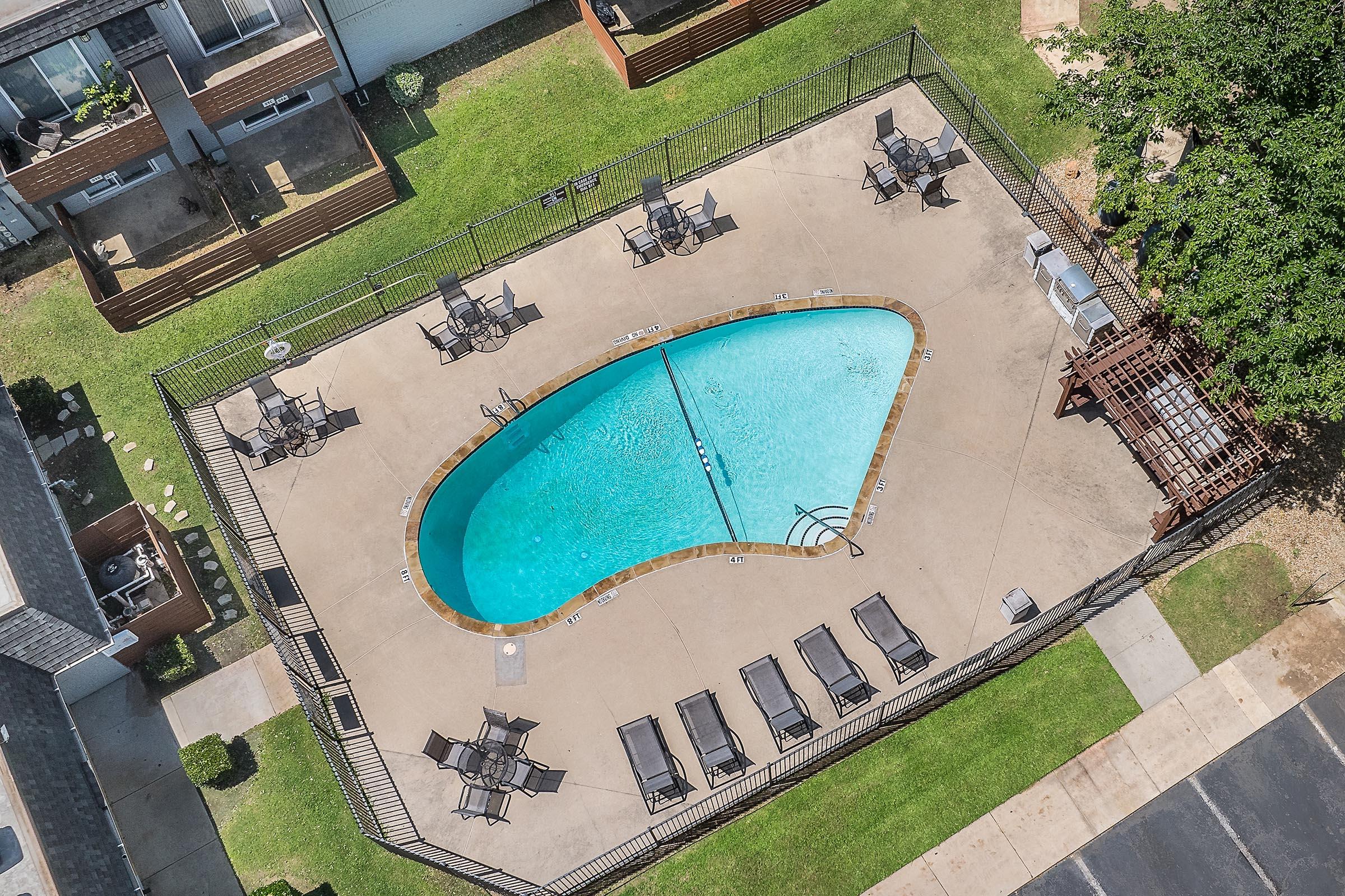 Aerial view of a residential swimming pool area. The pool has an irregular shape and is surrounded by a safety fence. Several lounge chairs are arranged around the pool on a concrete deck, with grass and nearby buildings visible in the background.