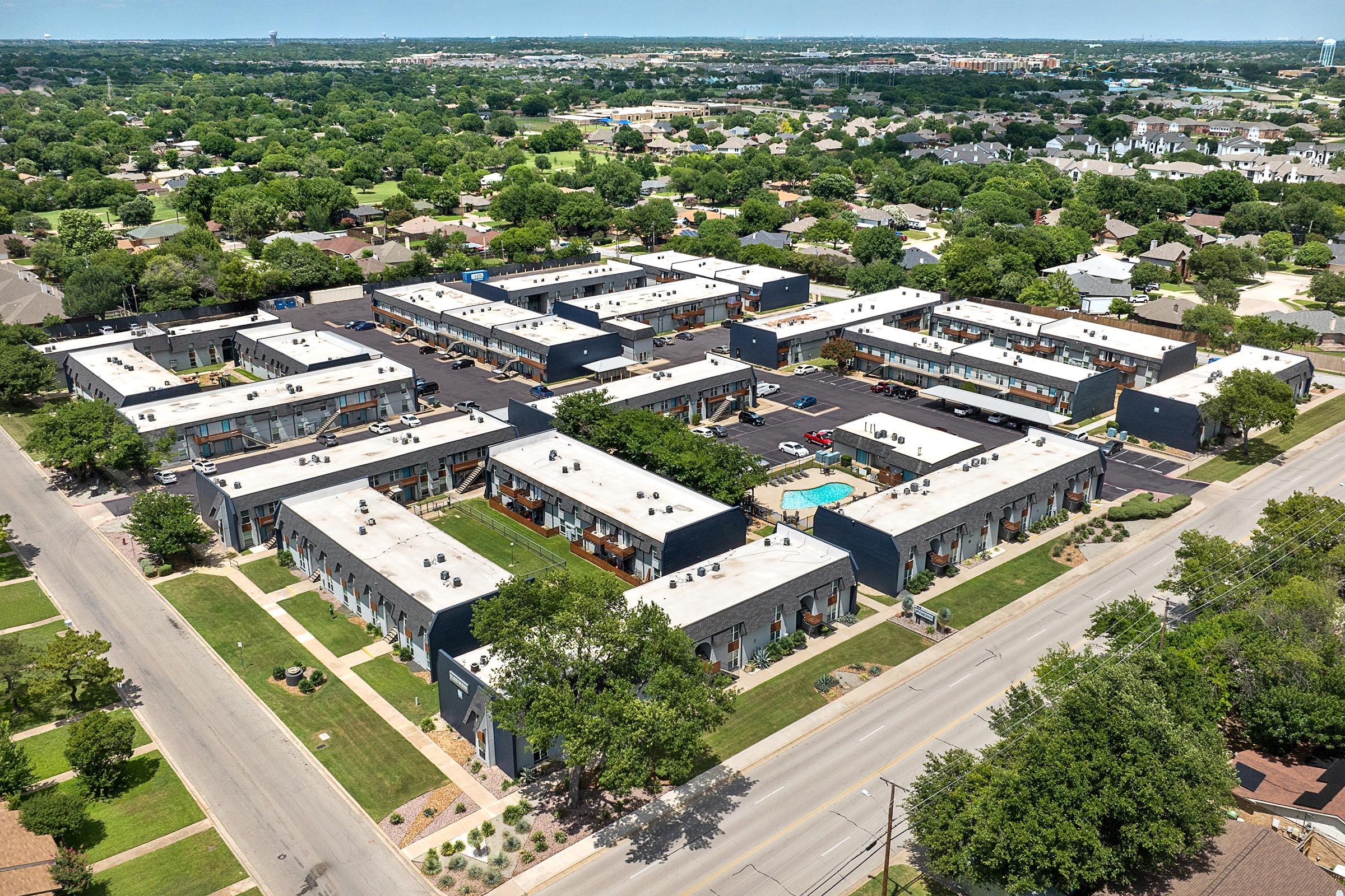 Aerial view of a residential complex featuring multiple buildings arranged in a grid pattern, with landscaped greenery and parking areas. A swimming pool is visible in the center, surrounded by well-maintained lawns. Nearby, single-family homes can be seen in a suburban setting.