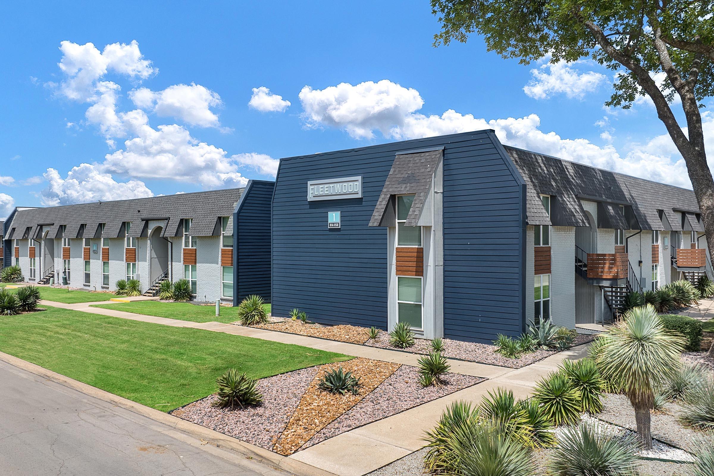 A view of a modern apartment complex named "Fleetwood," featuring two-story buildings with a mix of gray and white exteriors. The landscaping includes greenery and rocks, with a pathway leading to the entrance. The sky is bright blue with fluffy white clouds.