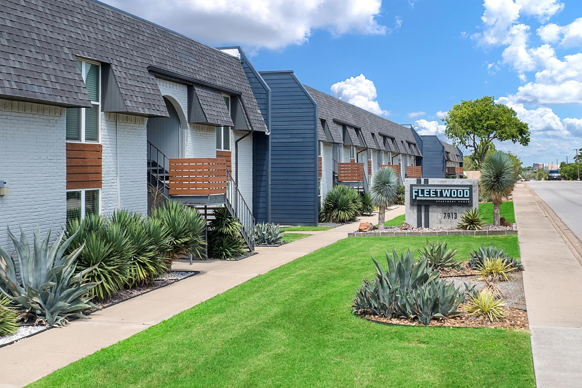 A view of an apartment complex called Fleetwood, showcasing a row of multi-story buildings with a mix of grey and white exteriors. There's a sign displaying the name and address, surrounded by well-maintained lawns and desert landscaping featuring agave plants. The sky is partly cloudy.