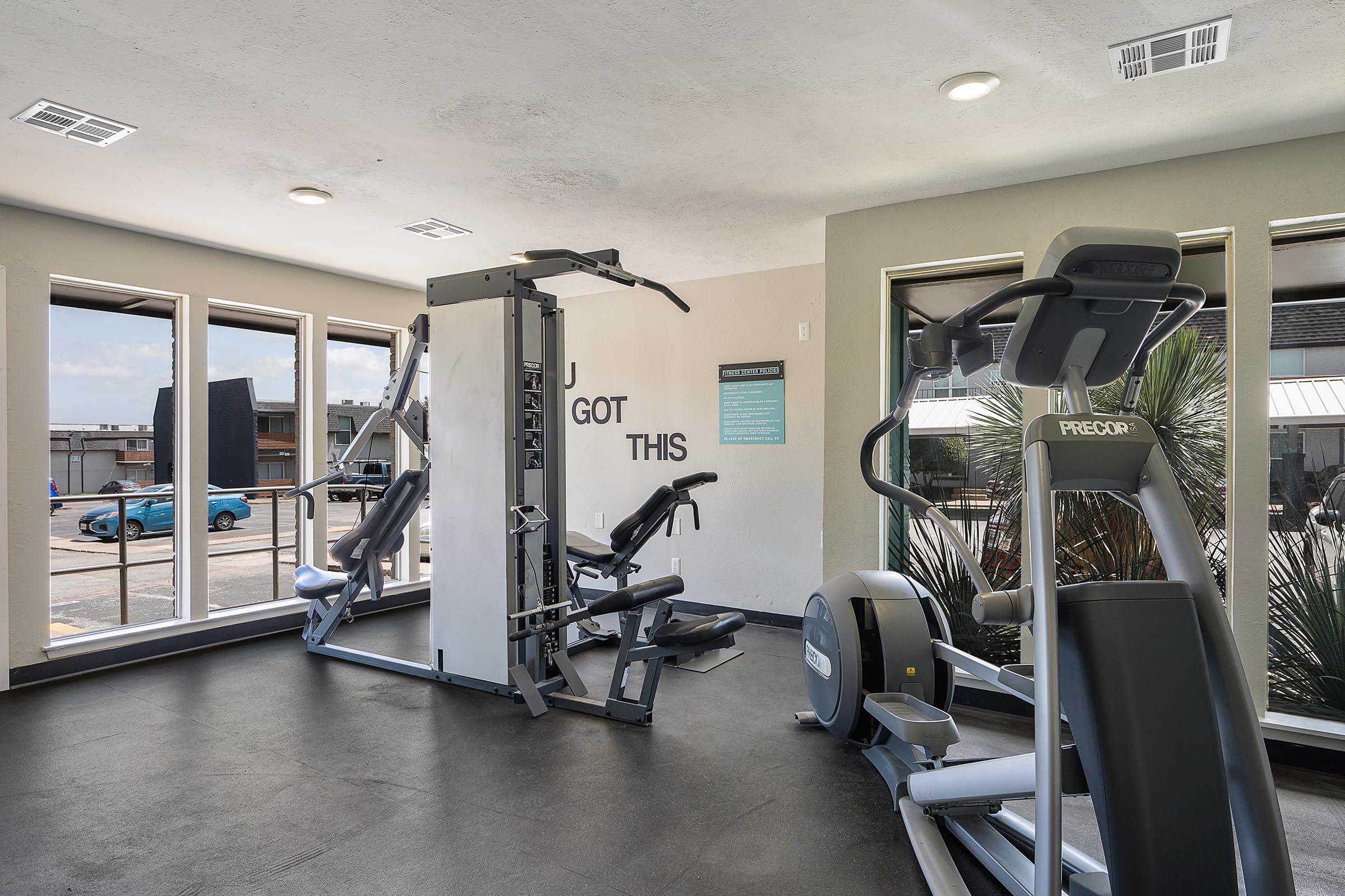 A well-lit gym featuring various exercise equipment, including a multi-gym machine, an elliptical machine, and a treadmill. Large windows allow natural light to fill the space, with a motivational message on the wall reading "YOU GOT THIS." A view of the outside is visible through the windows.