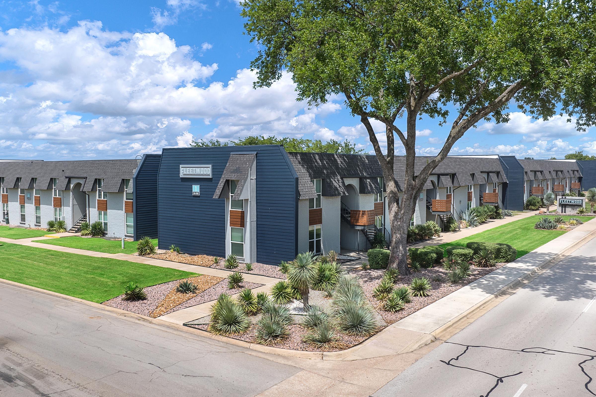 A modern apartment complex with a sleek design featuring a mix of dark blue and light-colored exteriors. The buildings are surrounded by well-maintained lawns and landscaping, including low shrubs and trees. A clear blue sky with scattered clouds is visible in the background.