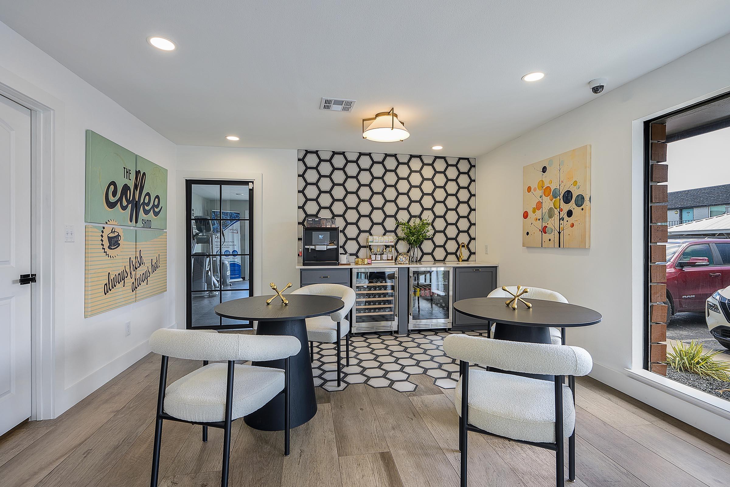 A modern coffee lounge featuring two round black tables with white chairs. The walls are decorated with abstract art and a coffee-themed sign. There is a beverage cooler on one side, and large windows allow natural light into the space, showcasing a view of parked cars outside. Stylish hexagonal tiles adorn the wall behind the cooler.