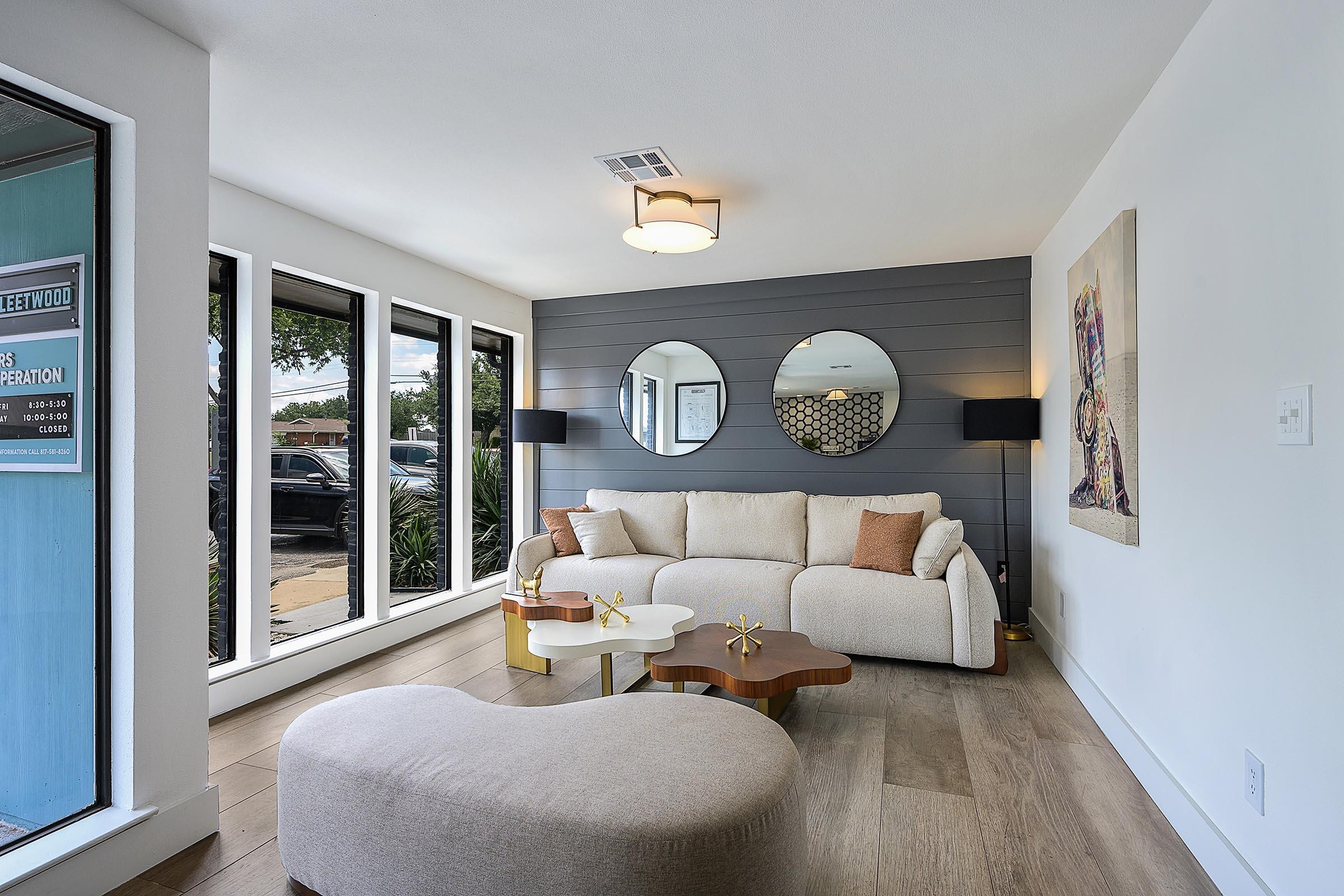 A modern living room featuring a light-colored sofa with orange accents, two round mirrors on the wall, a small white coffee table, and a unique curved lounge seat. Large windows let in natural light, showcasing a view of the outdoors. Stylish decor includes wall art and contemporary lighting.