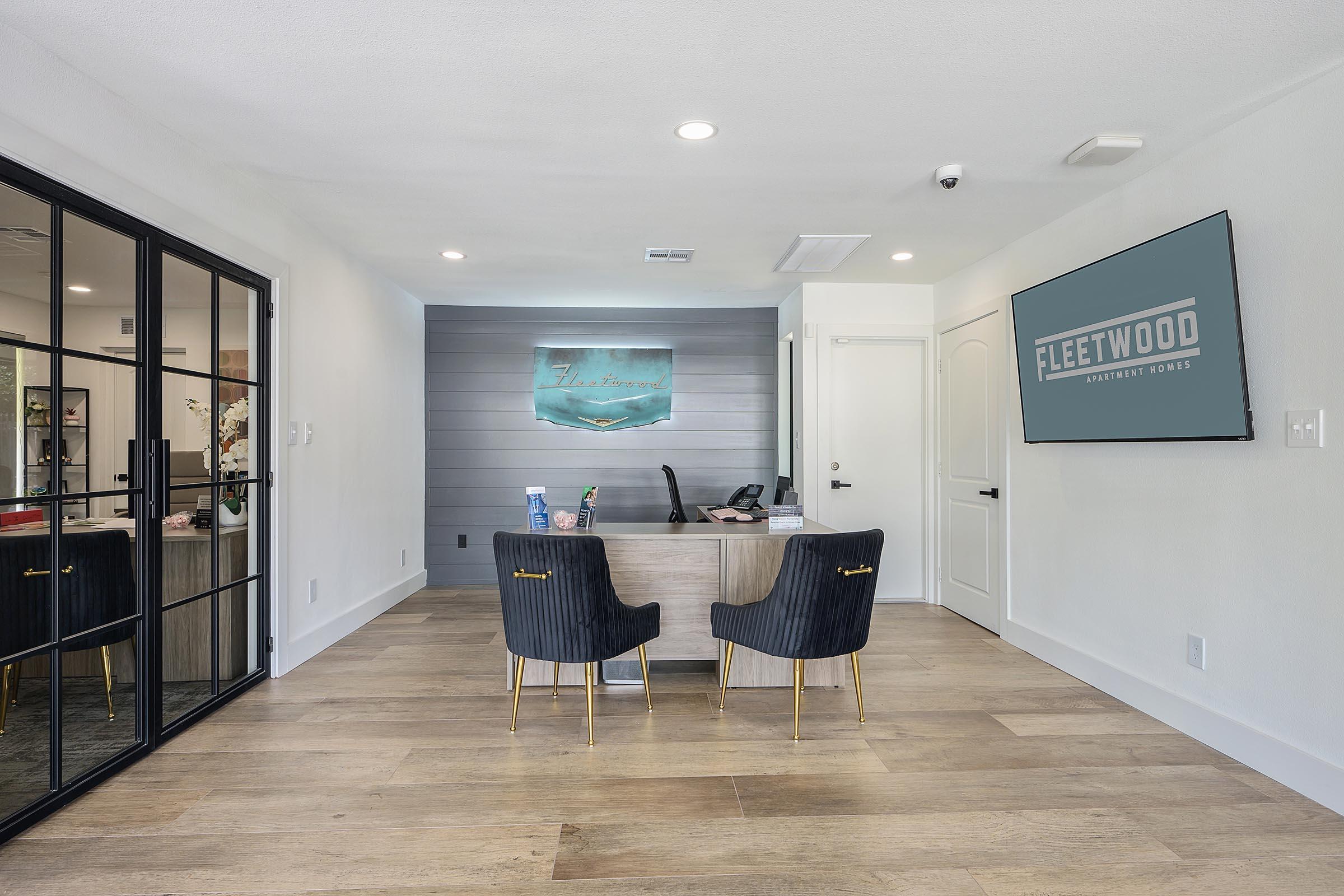 Interior view of a modern office reception area featuring a minimalist desk, two stylish black chairs, and a calm color palette. A wall sign reading "FLEETWOOD APARTMENT HOMES" is visible, along with a softly lit decorative piece on the adjacent wall. Natural light enhances the inviting atmosphere.