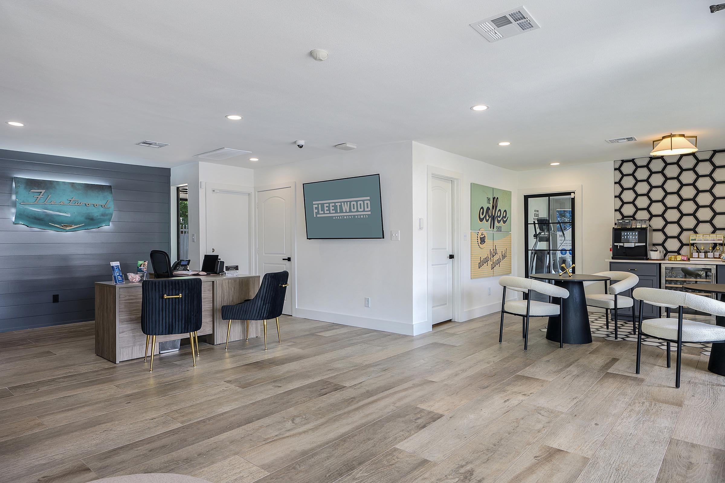 A modern reception area with a wooden front desk, two stylish chairs, and a digital screen displaying a graphic. There is a coffee station and a seating area with circular tables and beige chairs. The walls feature decor including the words "Fleetwood" and "Coffee," along with a textured panel design. Bright and welcoming ambiance.