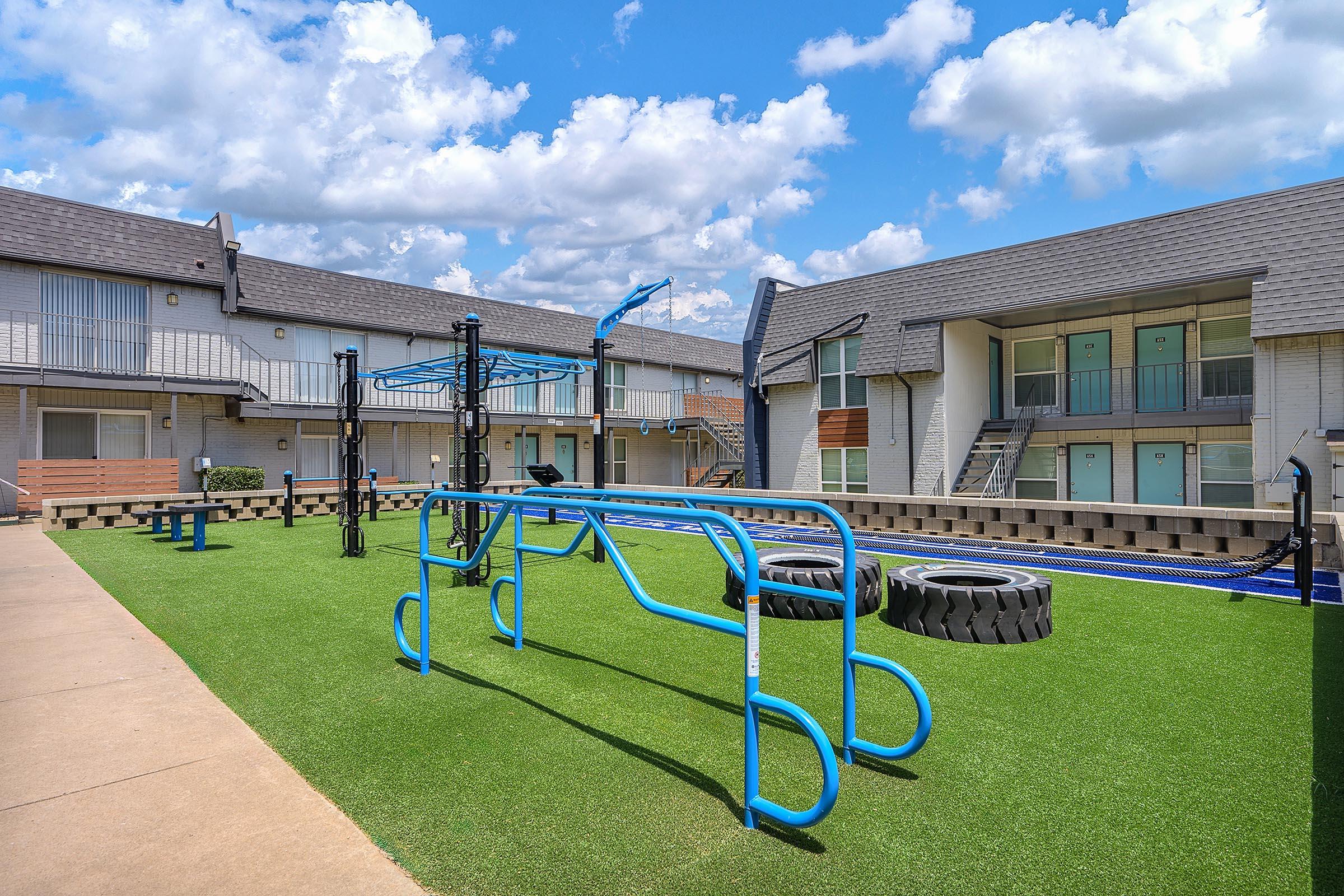 Playground area with blue exercise equipment and large tires on a green synthetic lawn. Surrounding the space are two-story apartment buildings with balconies and a blue sky filled with fluffy white clouds. The scene is inviting and designed for recreation and fitness.