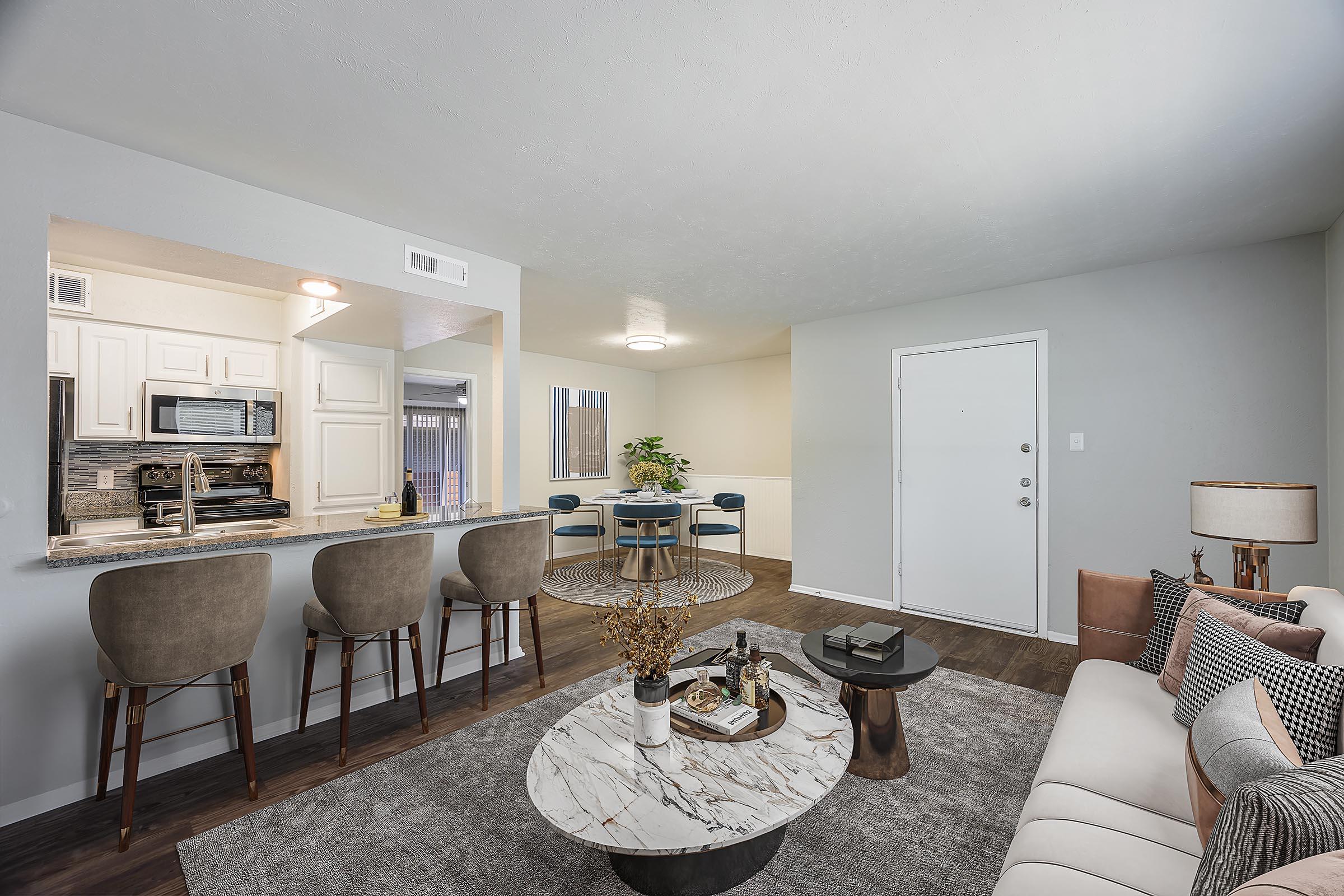 A modern apartment interior featuring a cozy living area. The space includes a beige sofa, a round marble coffee table, and a decorative rug. A kitchen bar with stools is visible, along with a dining area containing a circular table and chairs. Soft lighting enhances the inviting atmosphere.