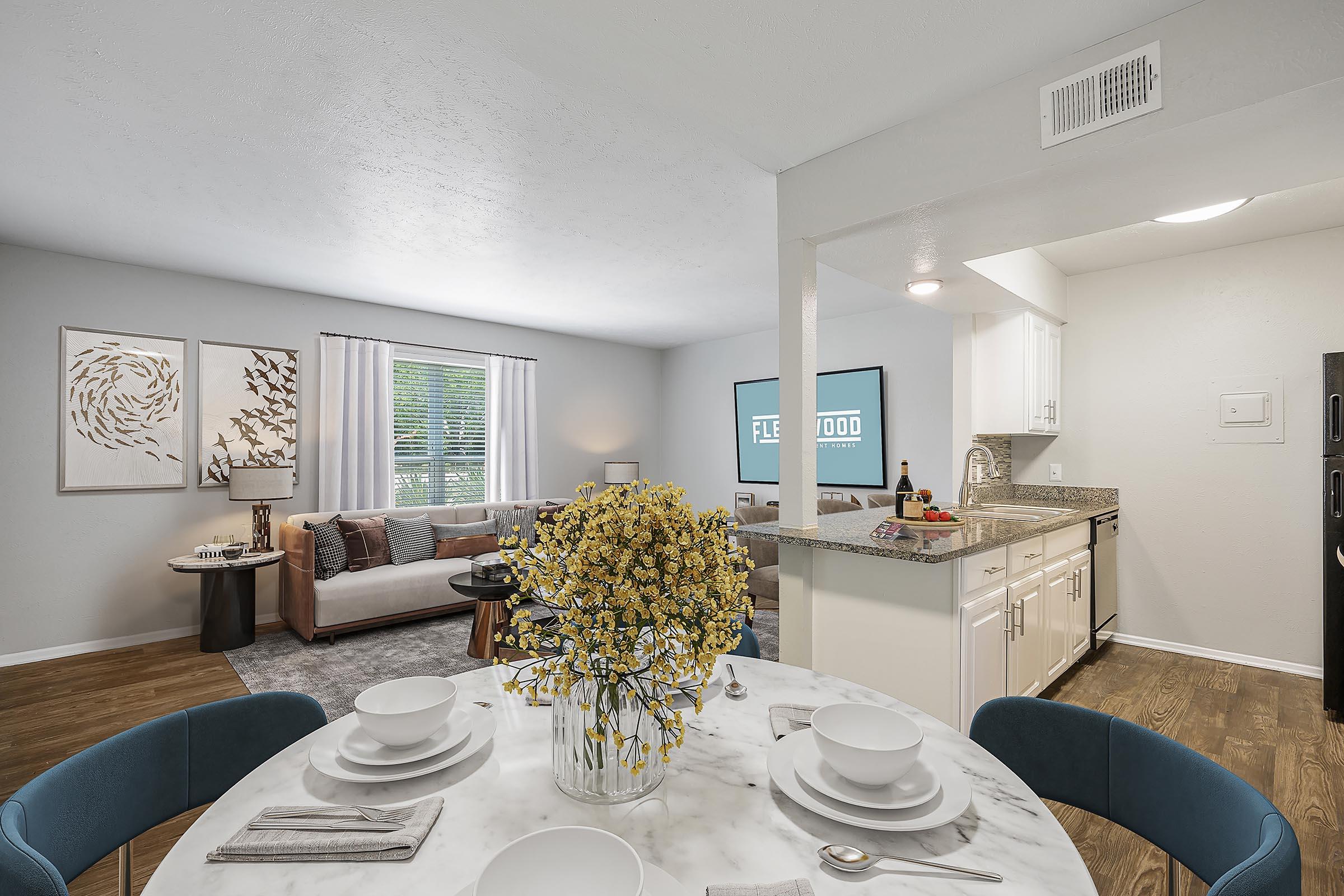 A modern, well-lit apartment interior featuring a dining table with white plates and a flower centerpiece, a cozy living area with a sofa and artwork on the walls, and an open kitchen with white cabinets. Natural light streams through large windows, enhancing the inviting atmosphere.