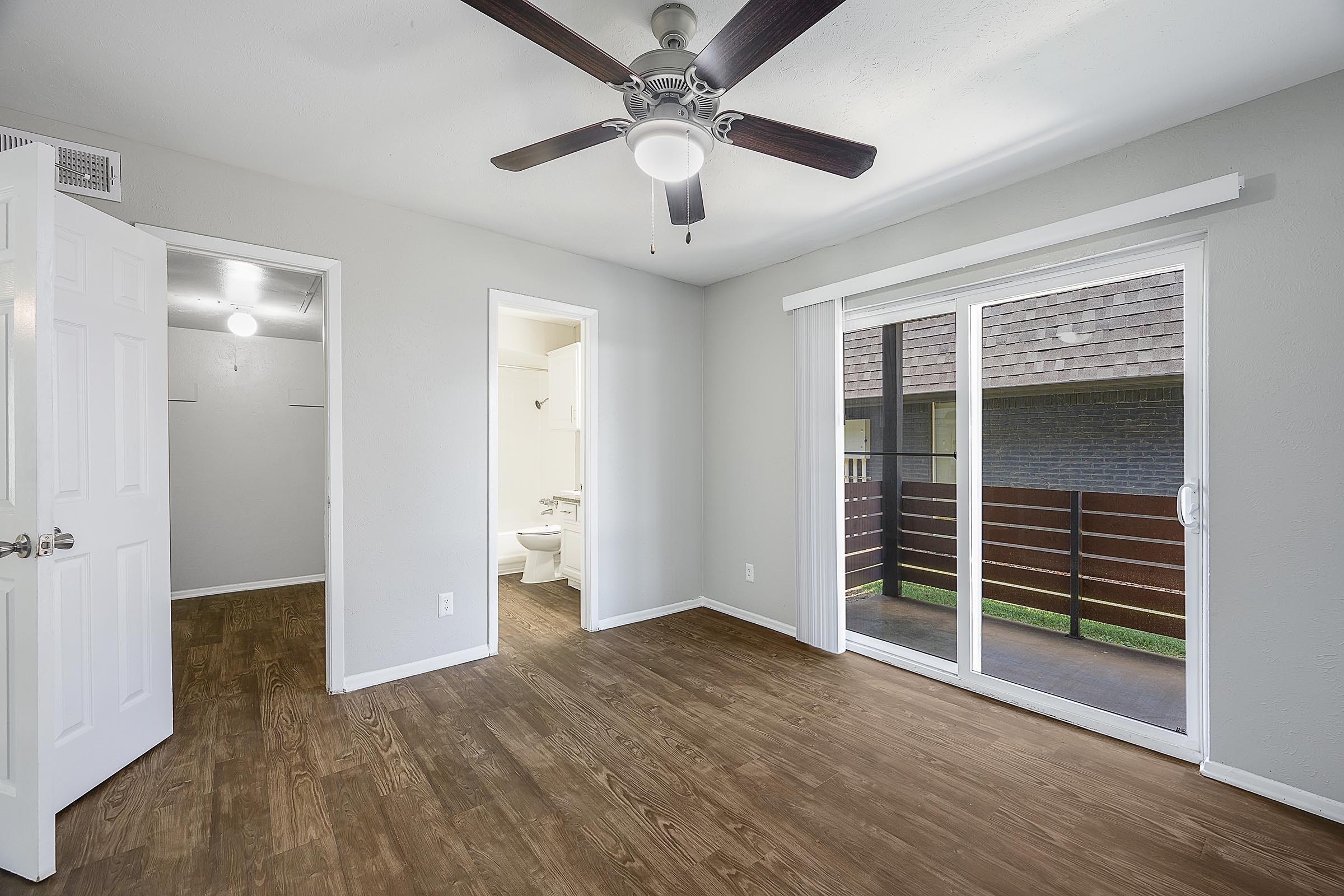 A spacious room with a ceiling fan and wooden flooring, featuring two open doors leading to a closet and a bathroom. A sliding glass door offers access to a balcony or patio area. The walls are painted in a light color, creating a bright and airy atmosphere.