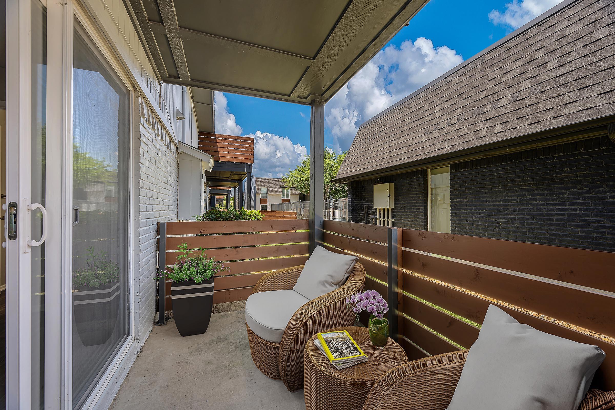 A cozy balcony with two wicker chairs, a small table, and potted plants. The space is surrounded by wooden fencing and features a view of a bright blue sky with clouds. Ideal for relaxation or enjoying a morning coffee.