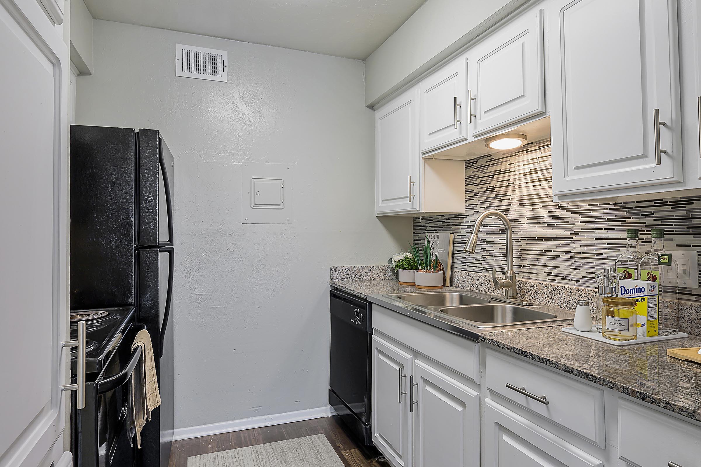 A modern kitchen featuring white cabinets, a granite countertop, and stainless steel appliances. A black refrigerator and dishwasher are visible. The backsplash has a mosaic design. A small plant sits near the sink, and a rug is placed on the wooden floor. Bright, neutral colors create a clean, inviting atmosphere.