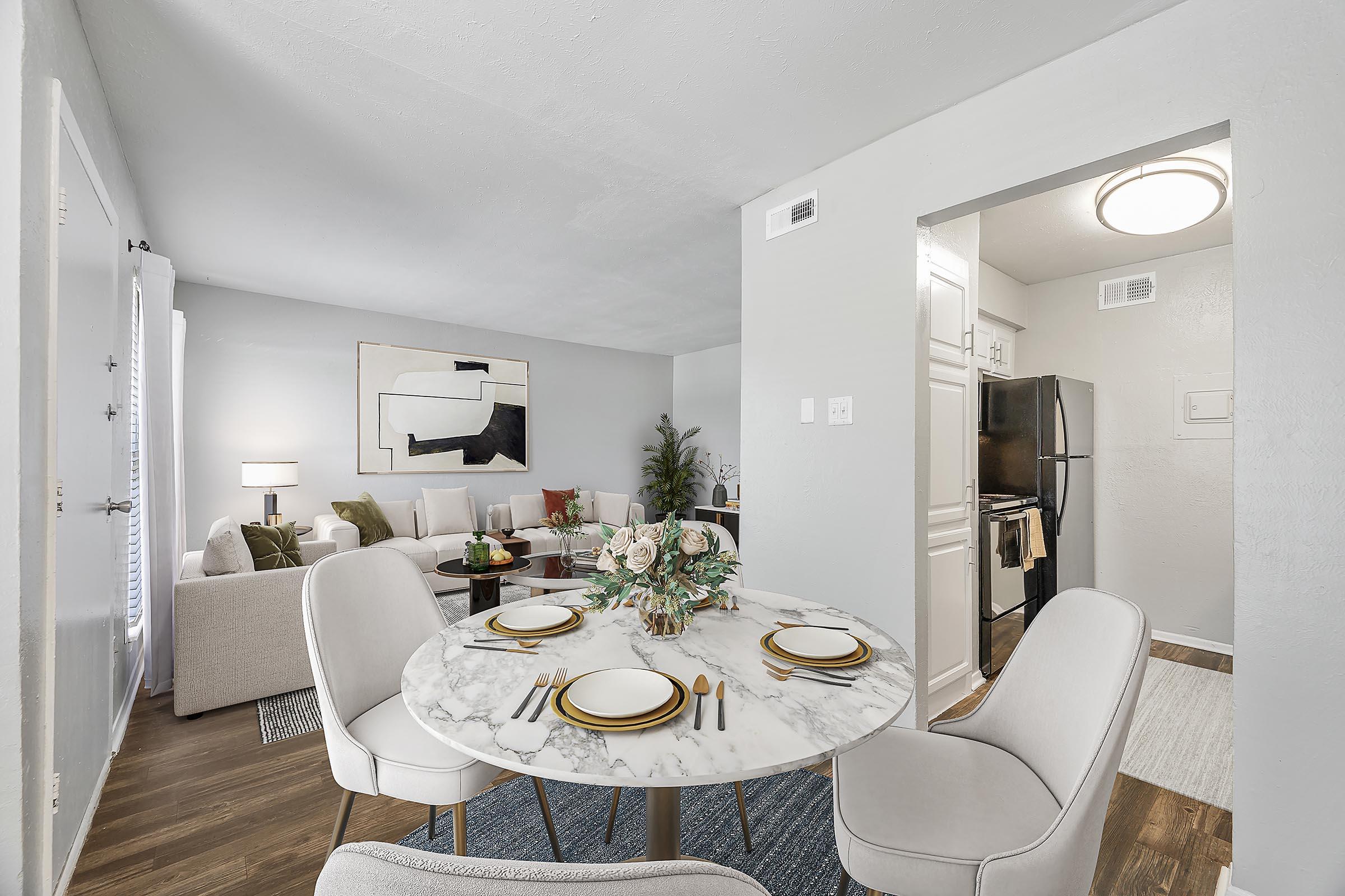 A modern dining area featuring a round marble table set for four with elegant plates and cutlery. The background shows a cozy living space with a light-colored sofa, decorative pillows, and greenery, complemented by a well-organized kitchen visible through an open doorway. The walls are painted in soft gray tones.