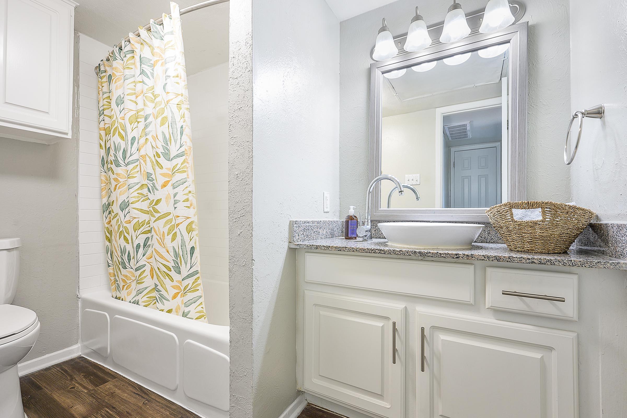 A well-lit bathroom featuring a tub with a floral shower curtain, a white cabinet with a sink, and a decorative mirror. The walls are painted in light gray, and there is a woven basket on the countertop. The flooring is dark wood, creating a warm contrast.