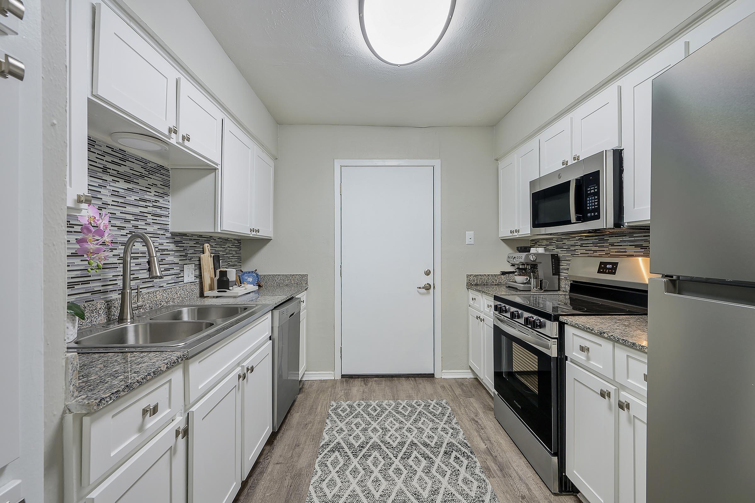 A modern kitchen featuring white cabinetry, granite countertops, stainless steel appliances (including a microwave, oven, and refrigerator), and a decorative backsplash. The space is well-lit with a ceiling fixture and has a light-colored door leading to another area. A gray area rug adds warmth to the floor.