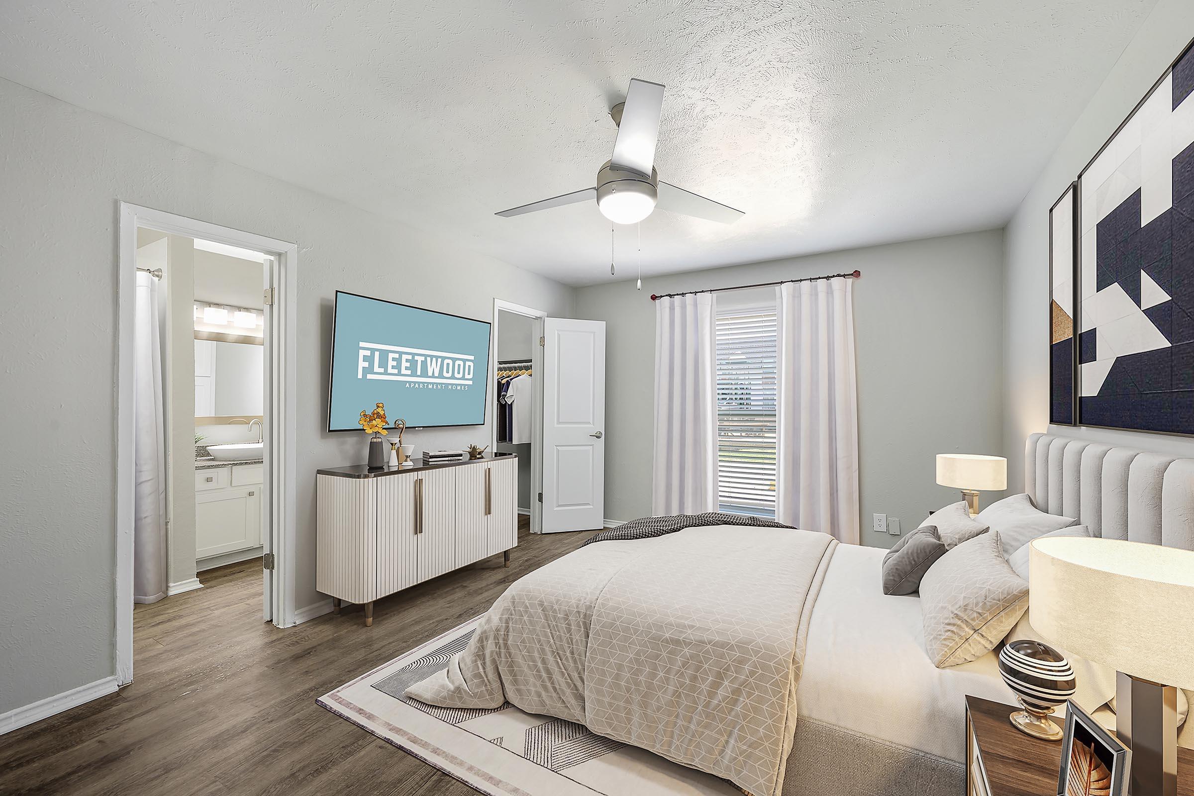 A cozy bedroom featuring a neatly made bed with light-colored bedding, a modern ceiling fan, and a stylish nightstand with a lamp. There’s a large flat-screen TV mounted on the wall displaying the "Fleetwood" logo. A door leads to a bathroom, and a window allows natural light, complemented by sheer curtains.