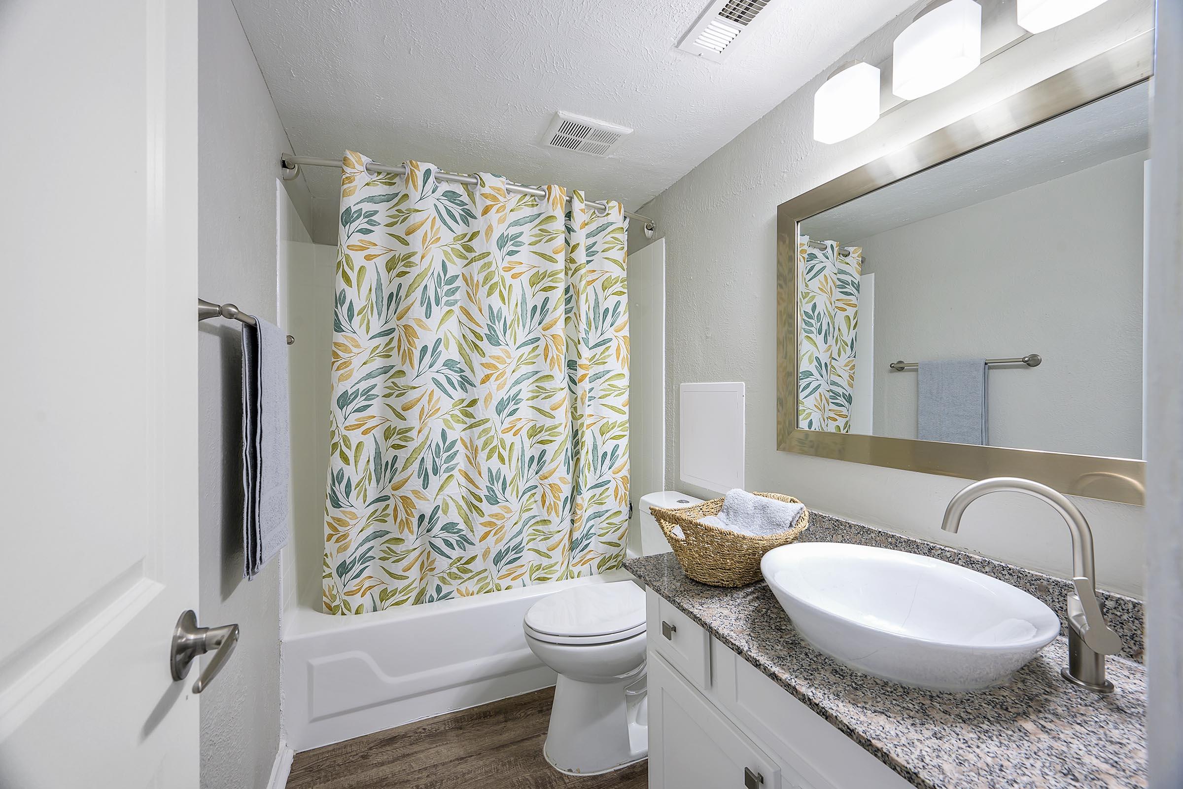 A bright, modern bathroom featuring a white bathtub with a leafy patterned shower curtain, a sleek countertop with a round sink, a mirrored cabinet above, and soft brown flooring. The space is well-lit with overhead lighting, and there are neatly arranged towels and a decorative basket on the counter.