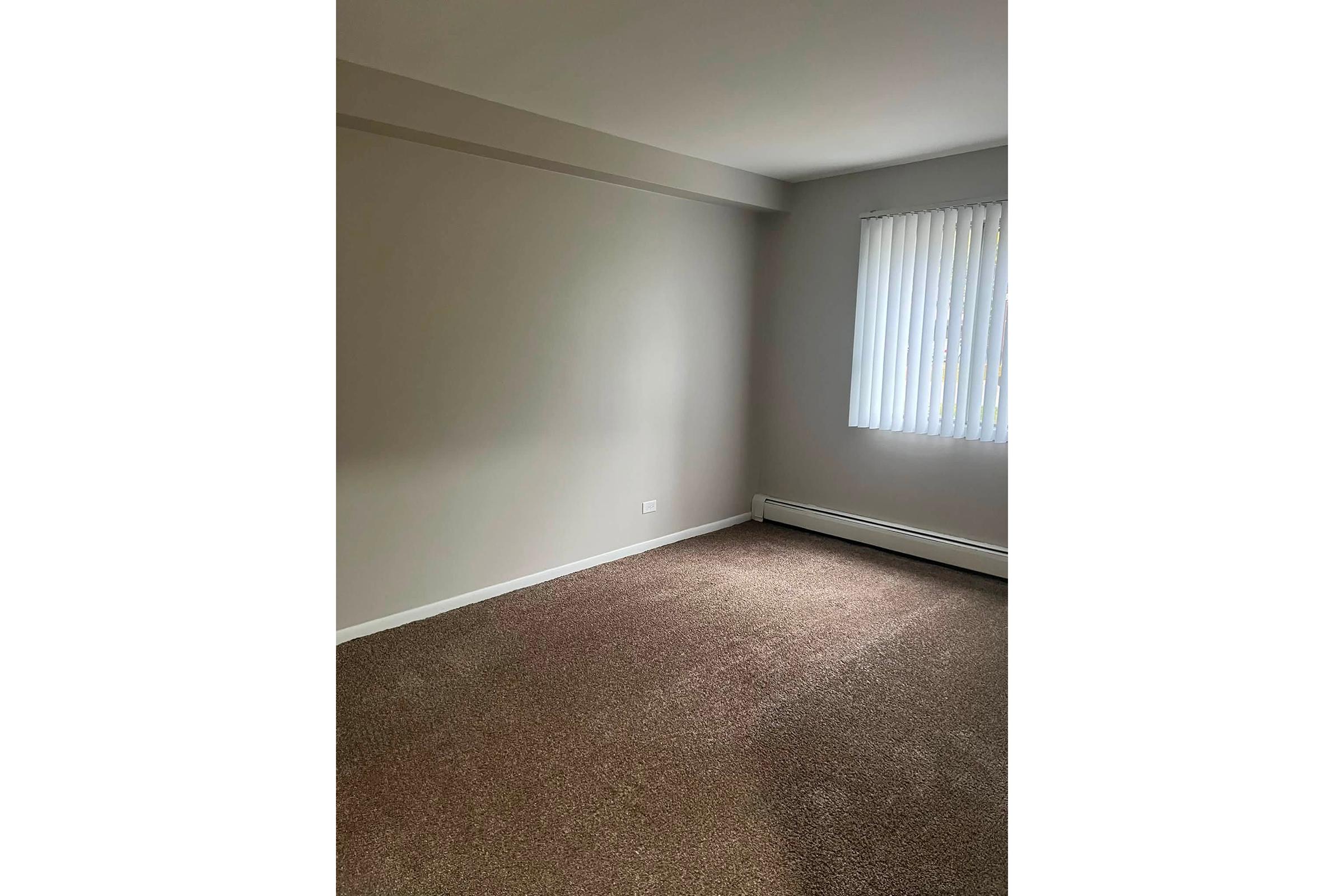 A vacant room with light brown carpet and beige walls. There is a window with vertical blinds letting in natural light. The room is sparsely furnished, giving it an empty appearance, with a radiator along one wall and no decorations or furniture visible.