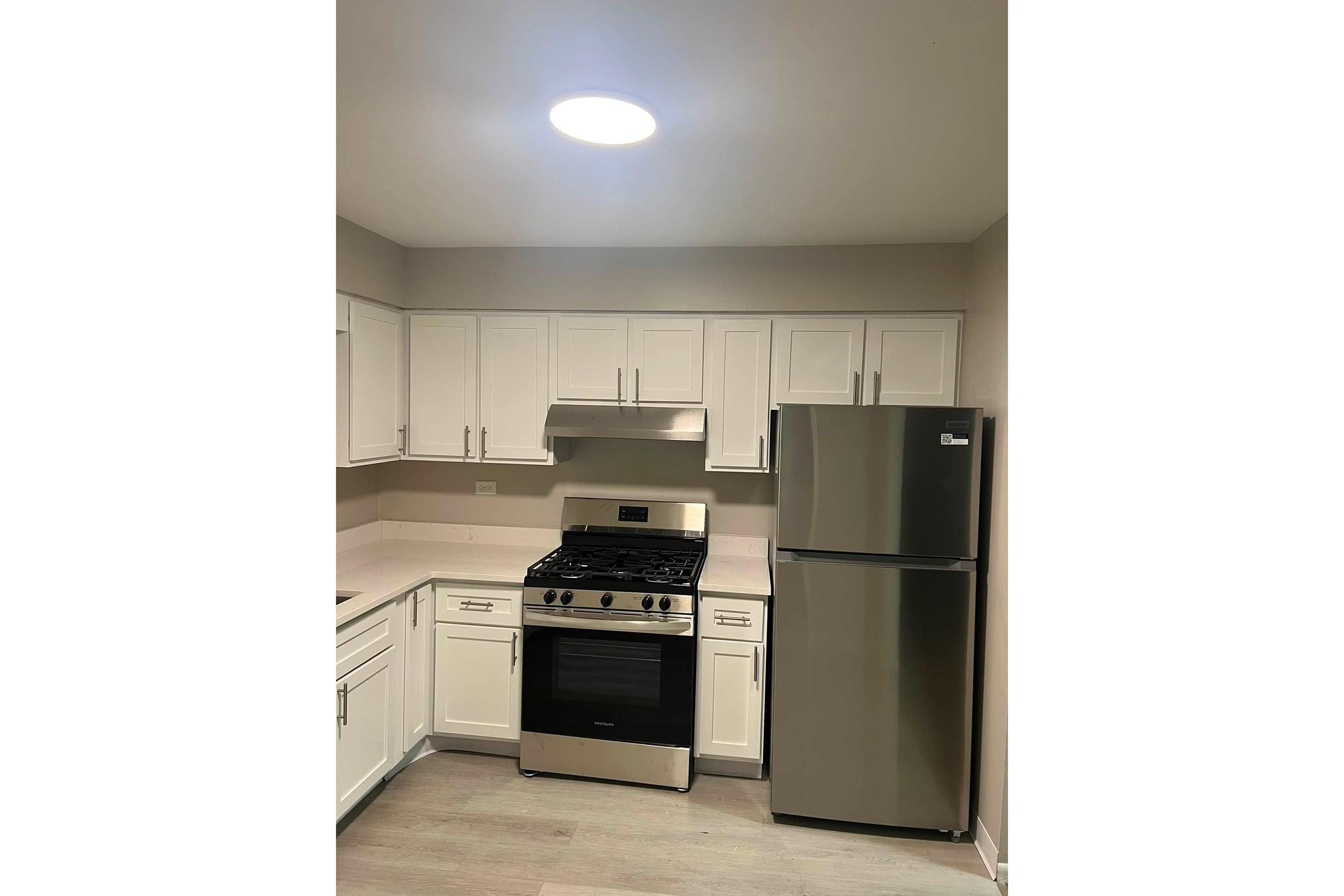 Modern kitchen with white cabinetry, a stainless steel refrigerator, and a gas range with a hood. The space is well-lit with a ceiling light, featuring a neutral-colored wall and light wood flooring, creating a bright and inviting atmosphere.