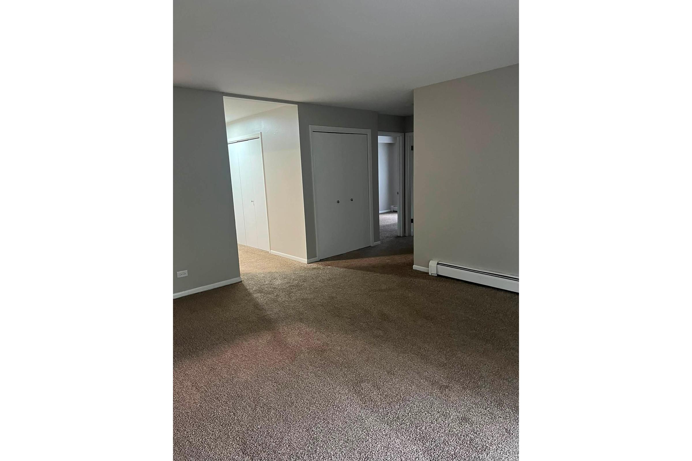 An empty room with beige carpet and gray walls. The space features doorways leading to other areas, including two closed closet doors on the left. Natural light enters from an unseen source, illuminating the room and highlighting its simplicity. The overall atmosphere is neutral and inviting.