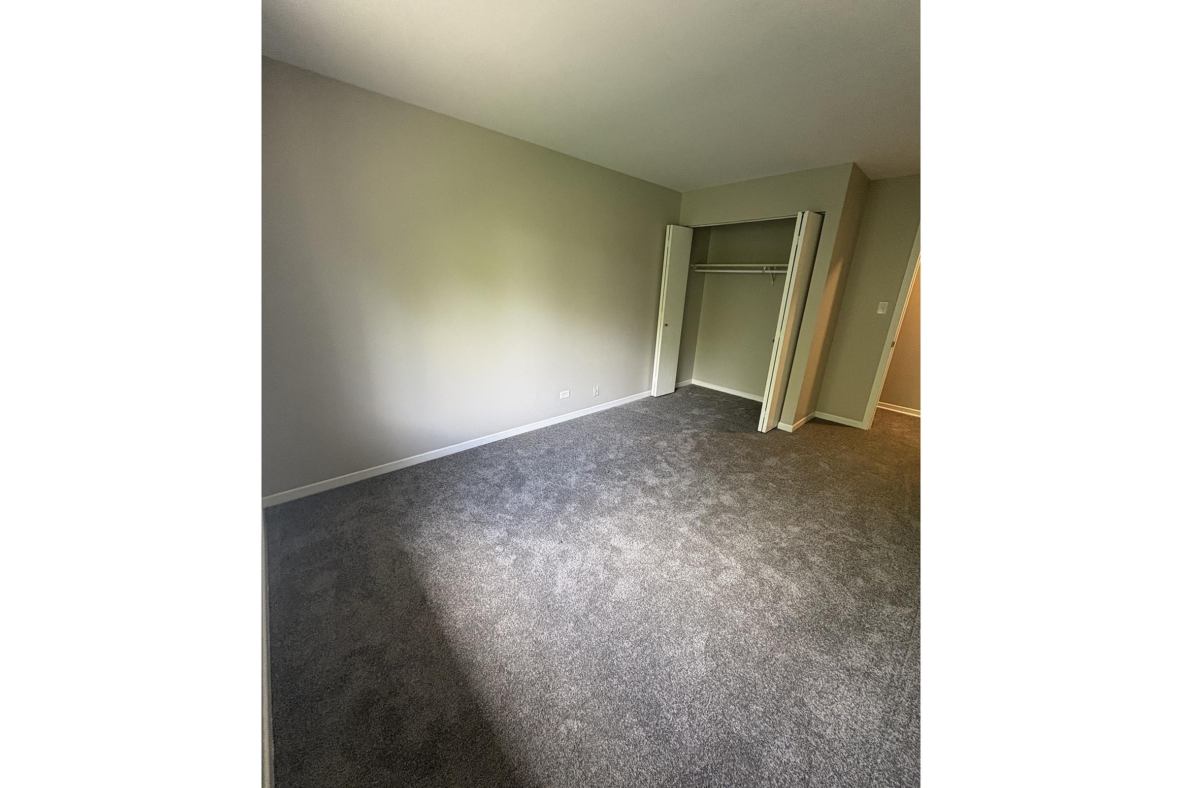 A spacious, empty bedroom with light gray carpet and neutral-colored walls. There is a closet with sliding doors on the left side of the image. Natural light is coming in from a window, creating a bright and airy atmosphere. The room appears clean and ready for furnishing.