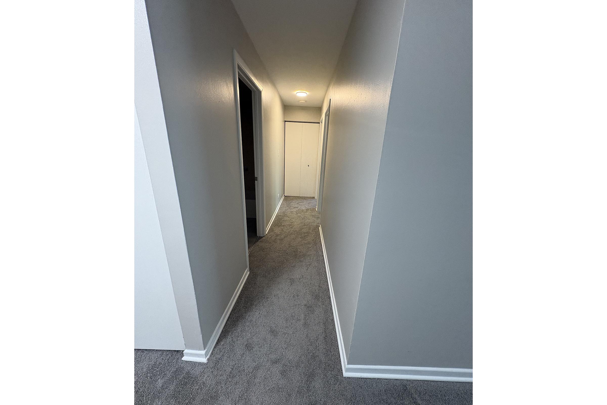 A well-lit hallway with light gray walls and newly installed carpet. The hallway has two doors on the left and a closed door at the end. Bright overhead lighting illuminates the space, creating a clean and modern look.