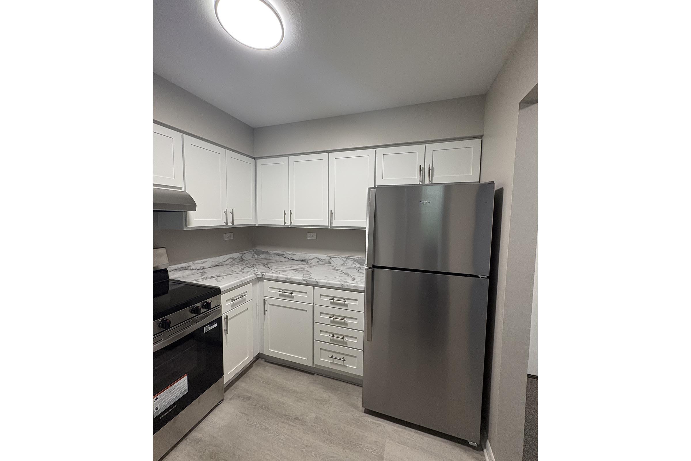 A modern kitchen featuring white cabinets, stainless steel refrigerator, and a marble countertop. The space is well-lit with a ceiling light, and there's a black stove with an oven. The flooring is a light color, complementing the overall contemporary design.