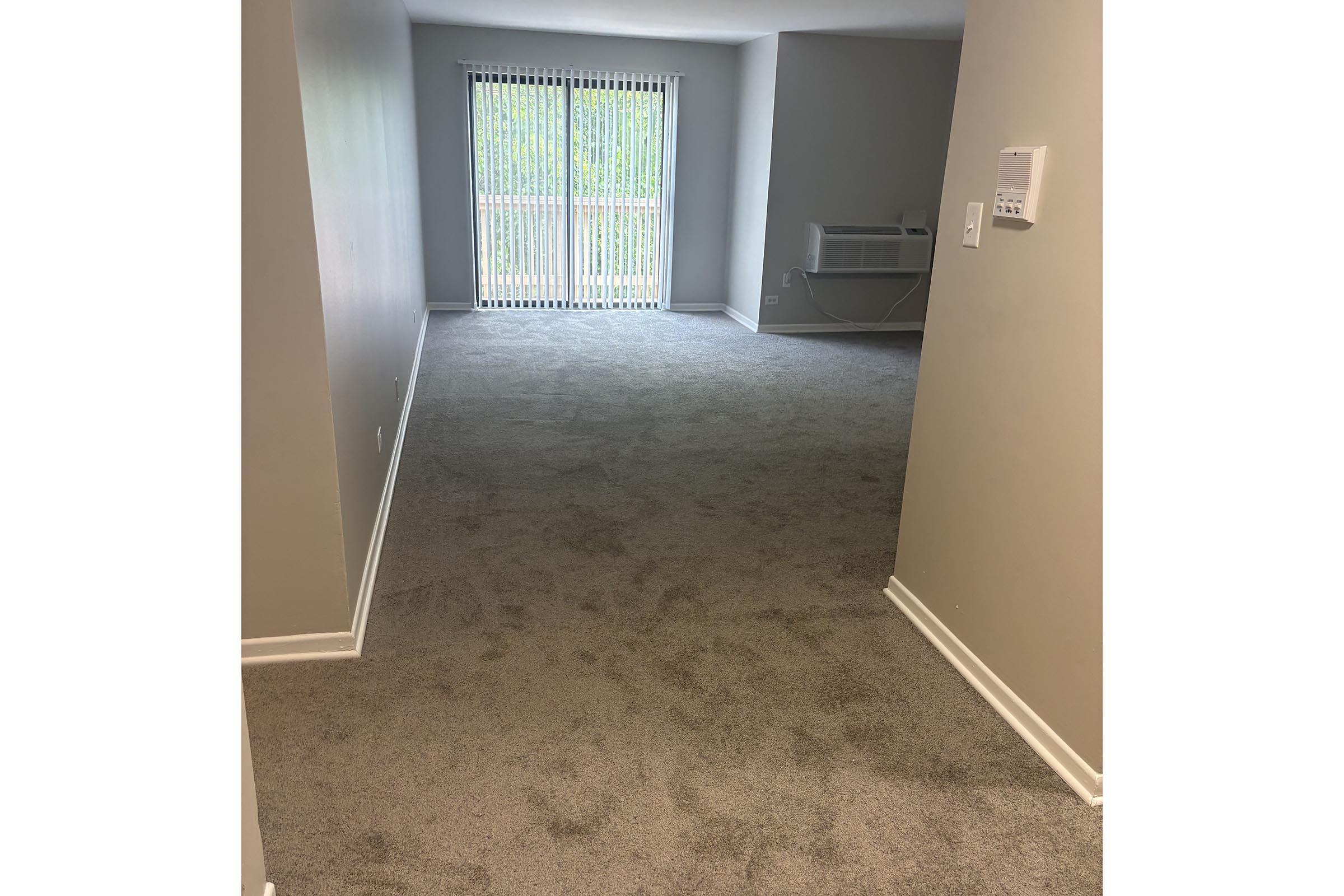 A spacious, empty room with light gray carpet and neutral-colored walls. There are vertical blinds covering a large window that leads to a balcony. An air conditioning unit is installed on the wall to the right. The overall ambiance is bright and inviting, showcasing a modern living space.