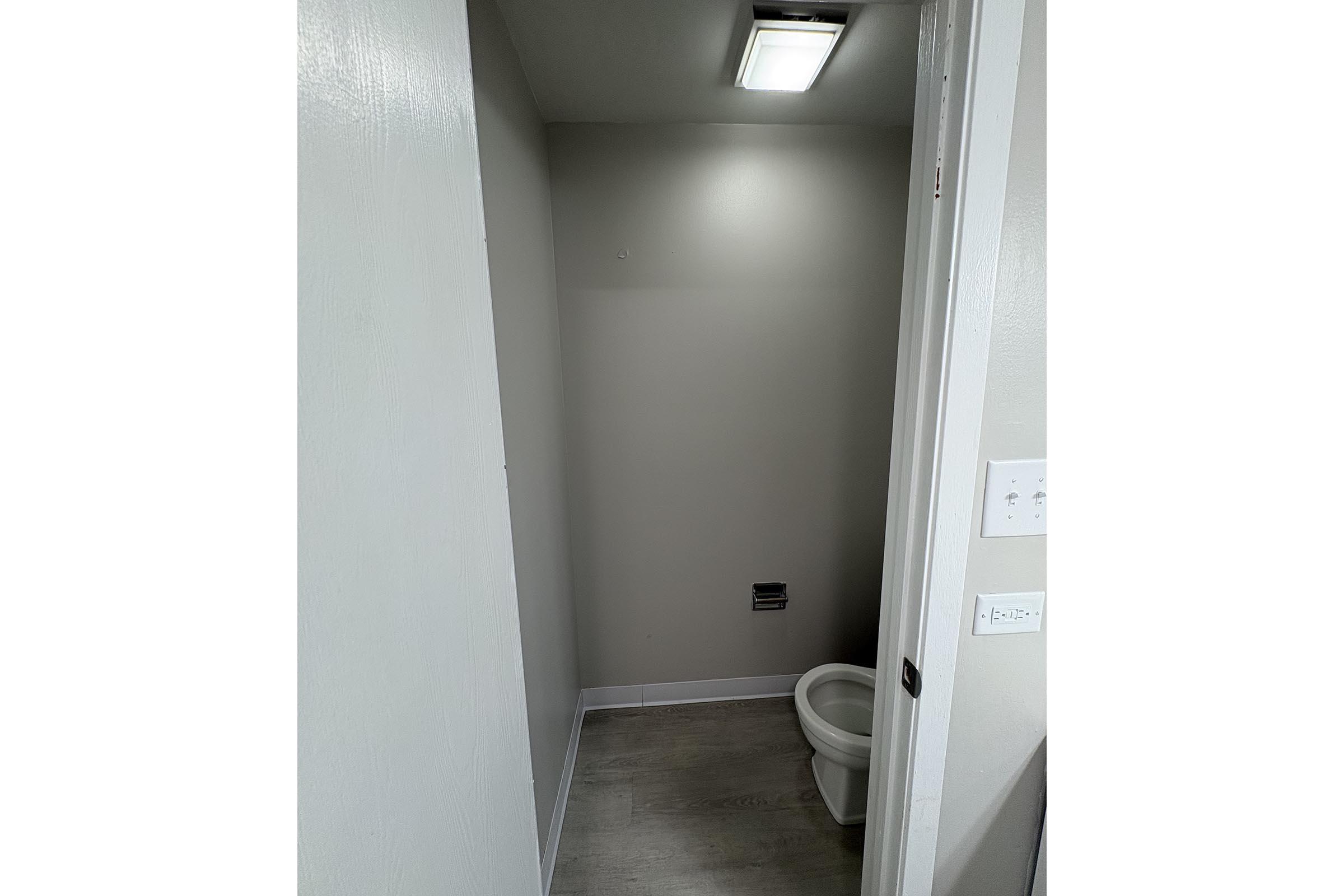 Small, minimal bathroom featuring a toilet positioned in a corner. The walls are painted a light color, and there's a ceiling light fixture overhead. A door is partially open, revealing the room's simple design with flooring in a light wood finish.