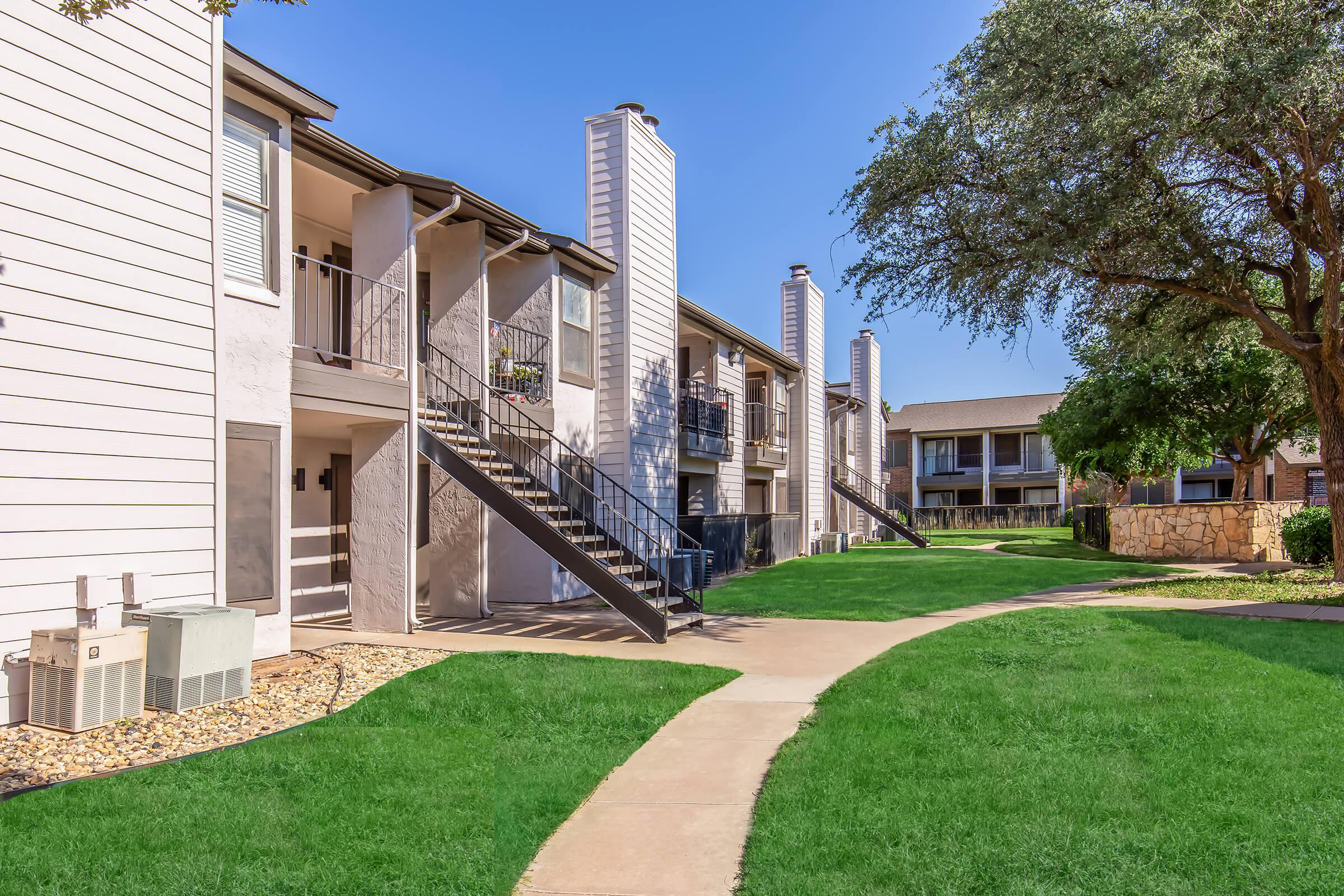 A view of an apartment complex featuring multiple two-story buildings. The exterior is characterized by light-colored siding, staircases leading to second-floor units, and neatly manicured green lawns. Paths connect the buildings, with some trees and landscaping visible in the background under a clear blue sky.