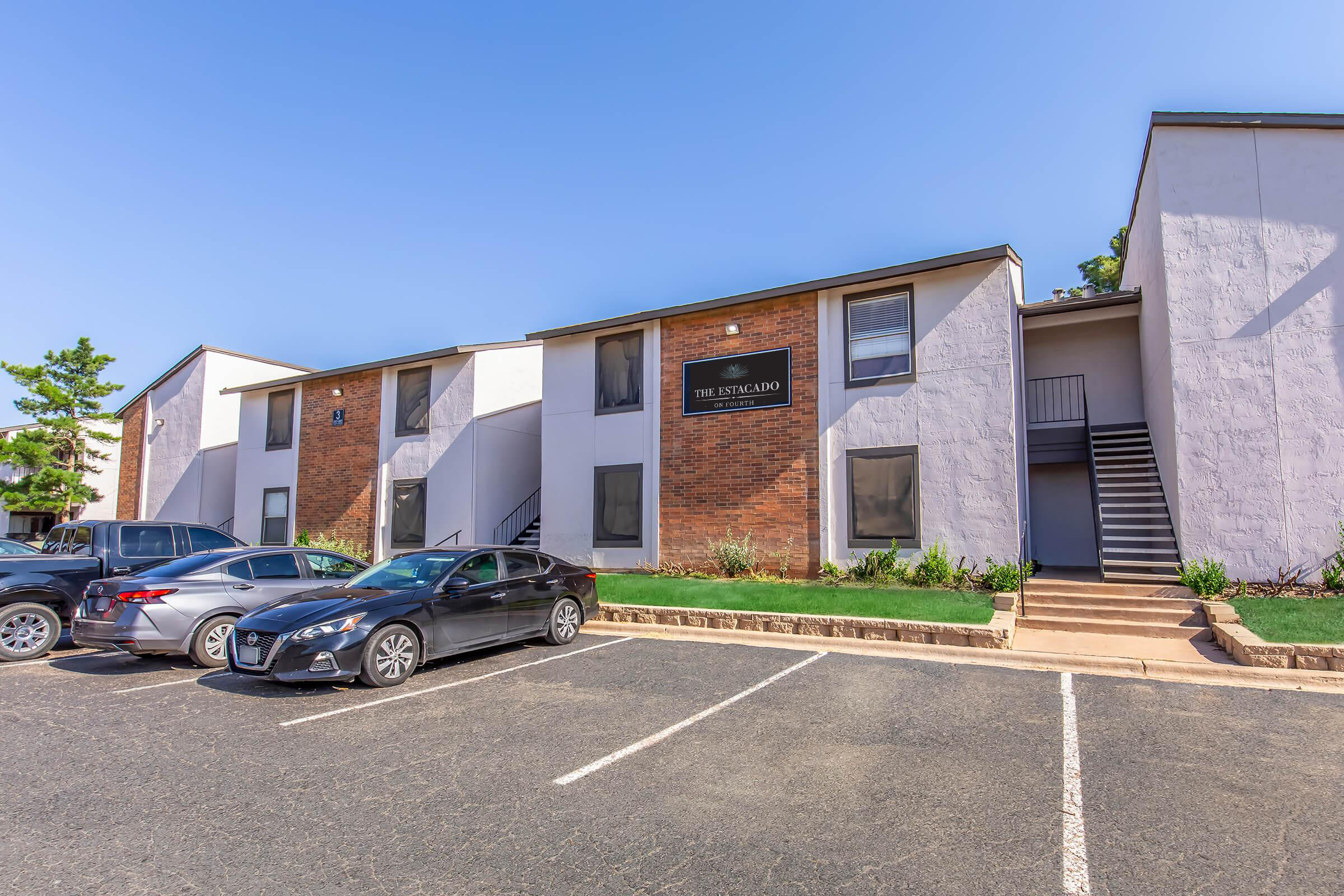 View of a multi-unit apartment building with a mix of brick and stucco exterior. The building features a sign reading "The Spectland." There are several cars parked in front, and a staircase leads to the upper level. The surrounding area includes grass and trees under a clear blue sky.