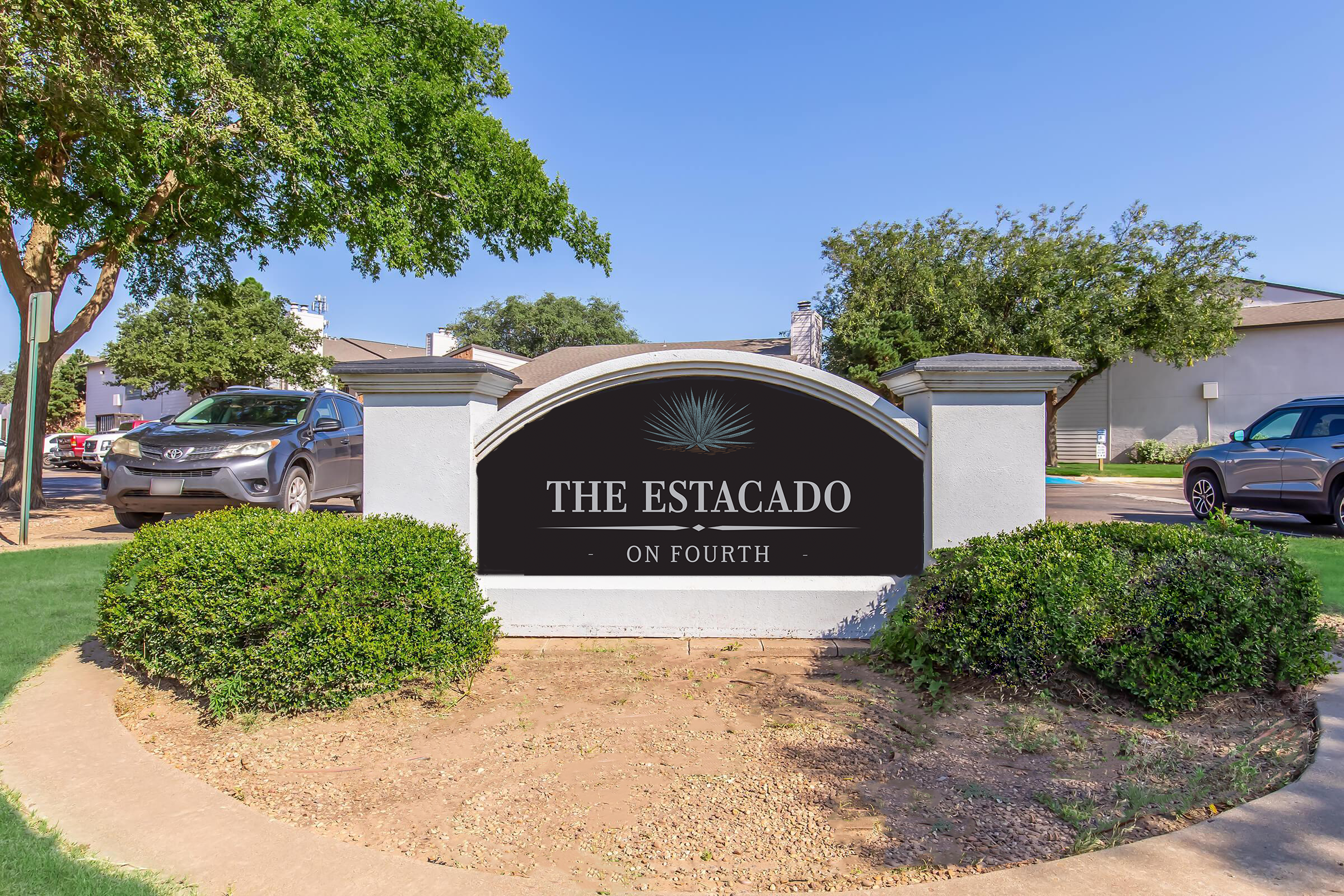 Sign featuring the text "The Estacado on Fourth," surrounded by a landscaped area with grass and shrubs. In the background, there are parked cars and a clear blue sky.