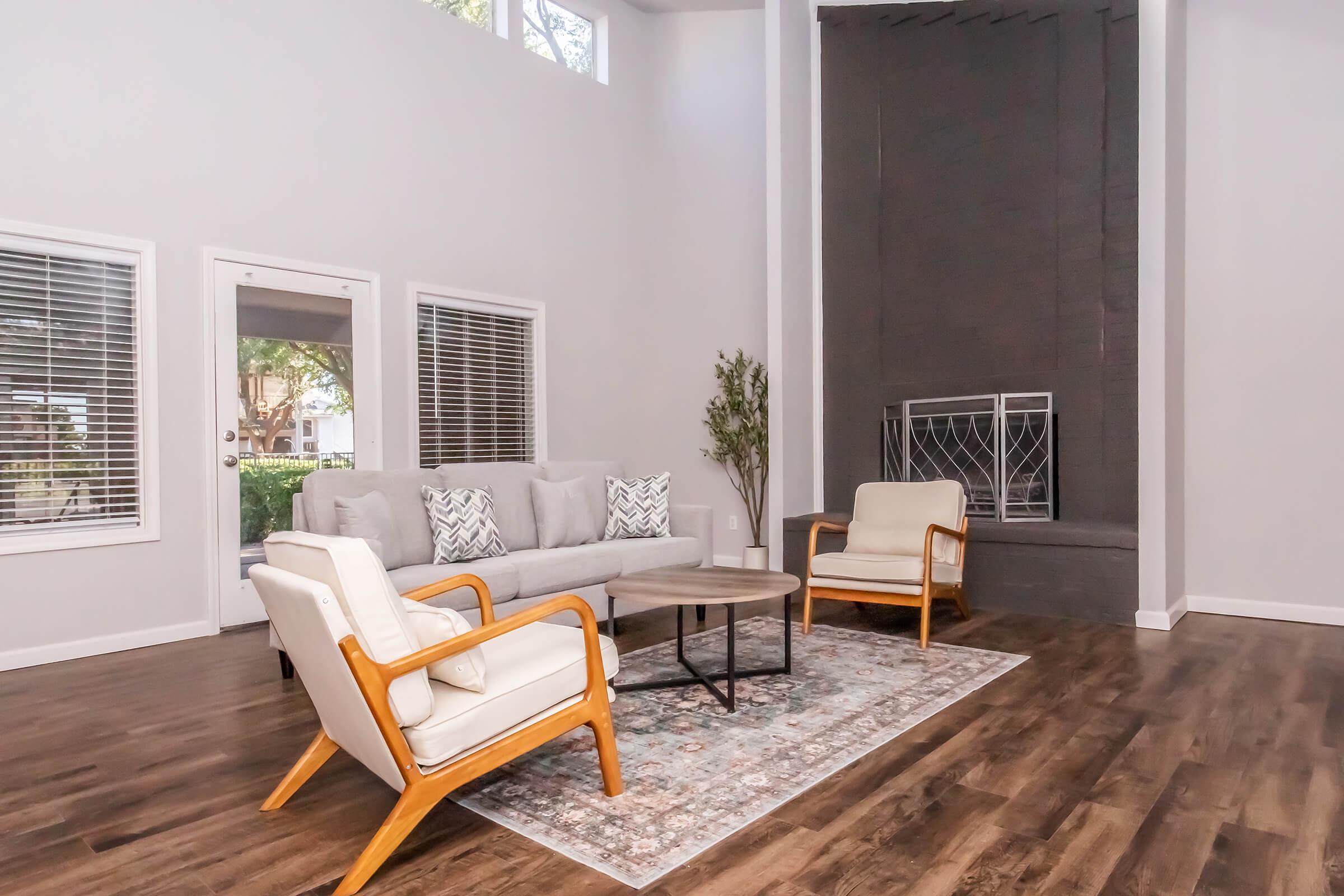 A spacious living room featuring a light gray couch with decorative pillows, two stylish armchairs, and a round coffee table on a patterned rug. Large windows allow natural light, and a modern fireplace is visible on the wall. The walls are painted in a soft gray tone, enhancing the contemporary feel of the space.