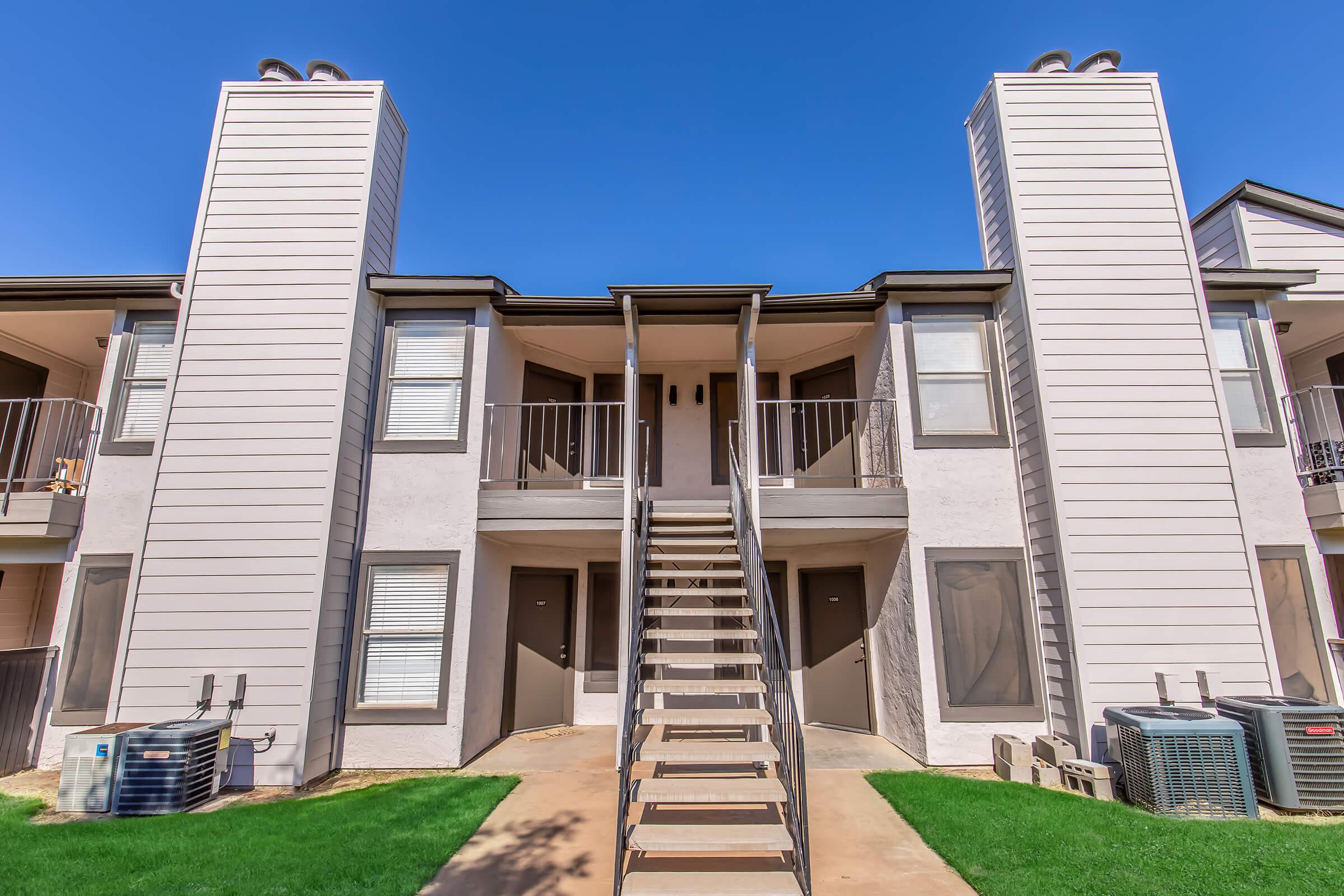 A two-story apartment building with two separate staircases leading to the second floor. Each unit features a balcony and windows on both levels. Air conditioning units are visible on the ground level, with landscaped green grass in the foreground and a clear blue sky above.