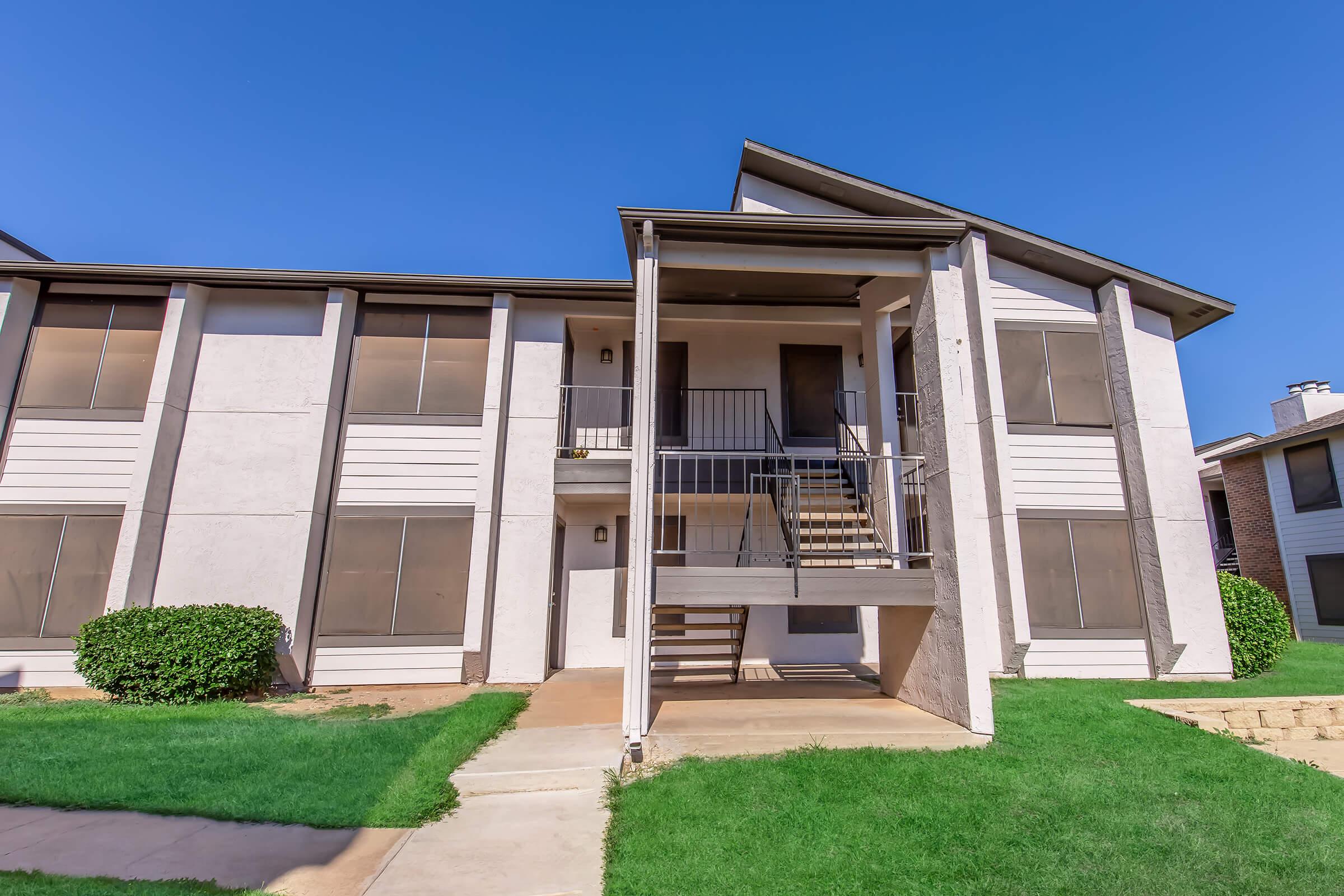 Exterior view of a two-story apartment building with a covered entrance and a staircase leading to the upper level. The building features large windows and is surrounded by green grass and small shrubs. The sky above is clear and blue.