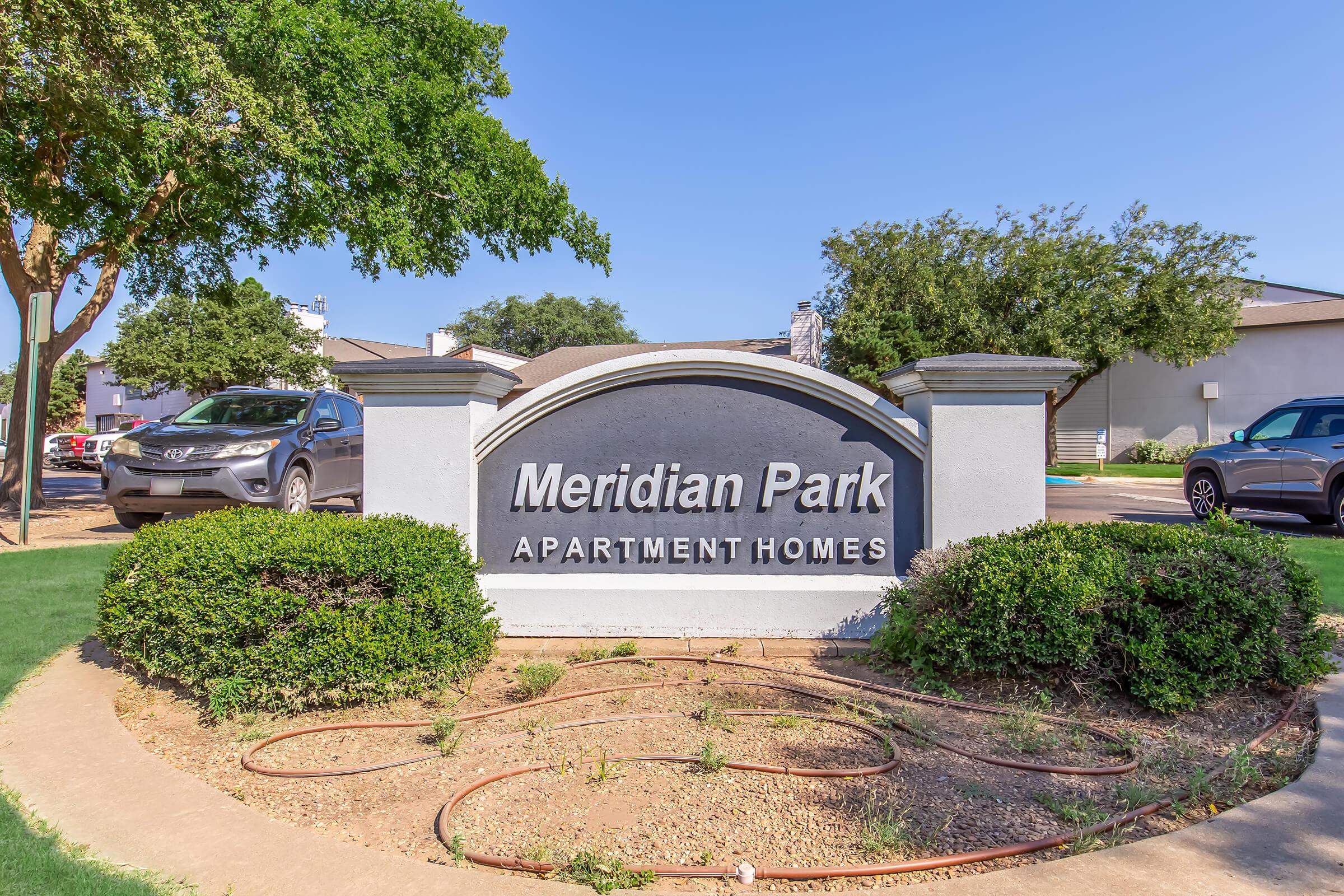 Sign for Meridian Park Apartment Homes with a well-maintained landscape featuring bushes and a clear blue sky in the background. Vehicles parked nearby. The sign is prominently displayed in an inviting manner.