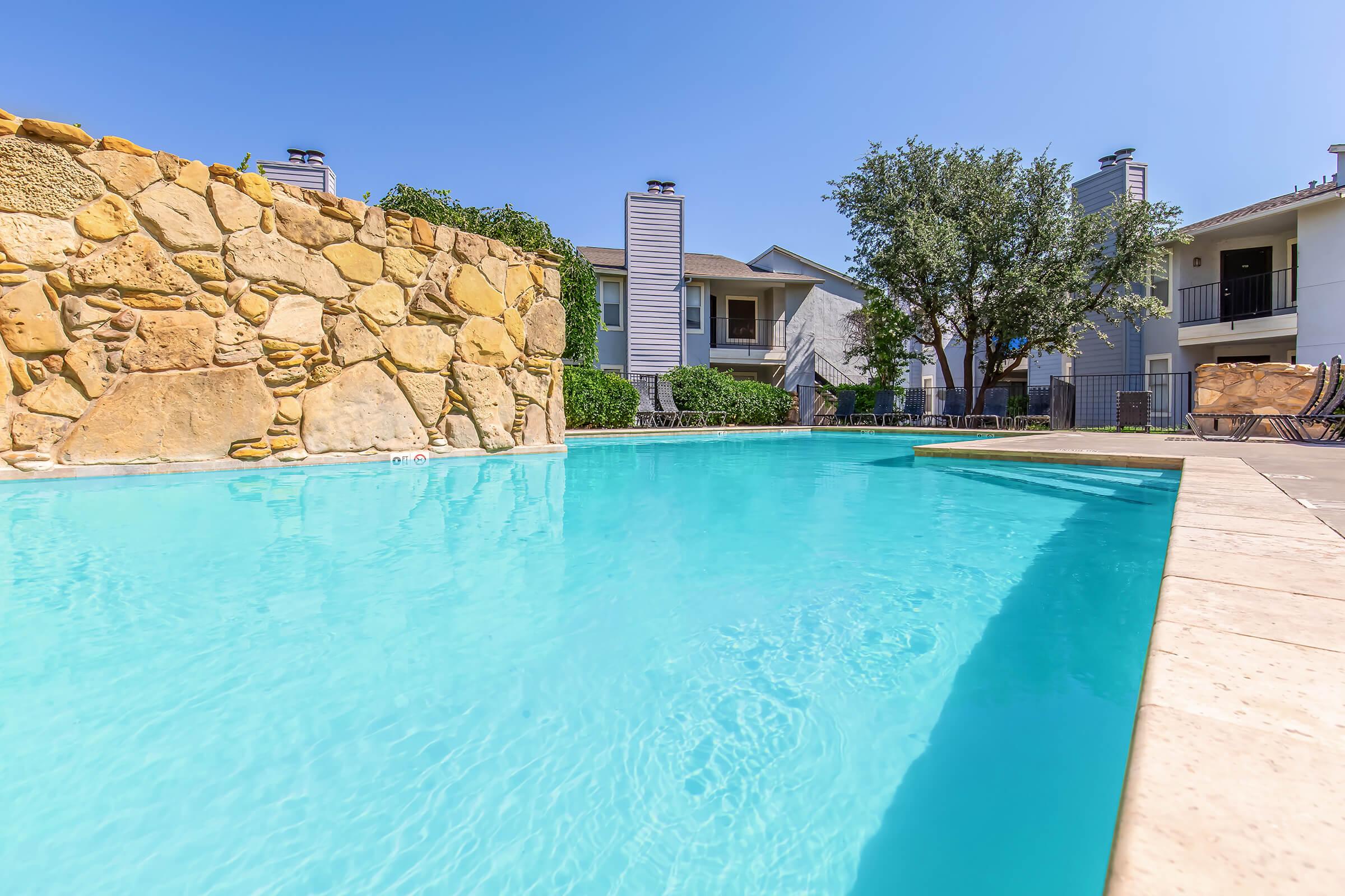 A clear blue swimming pool with a stone wall on one side, surrounded by lush greenery and residential buildings in the background. The sky is bright and sunny, creating a relaxing atmosphere.