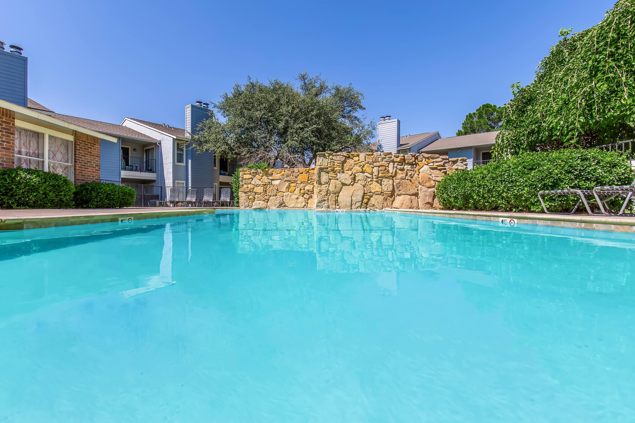 A clear blue swimming pool surrounded by green shrubs and a stone wall, with nearby apartment buildings and a clear blue sky in the background. The scene conveys a serene and inviting atmosphere, perfect for relaxation.