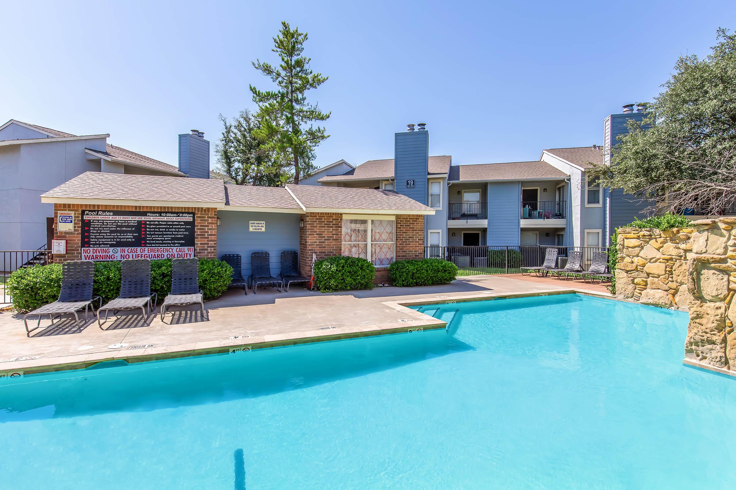 A clear blue swimming pool surrounded by lounge chairs, with a brick building and multiple apartments in the background. Lush green bushes line the pool area, and a warning sign is posted near the pool. The scene is set under a bright, sunny sky.