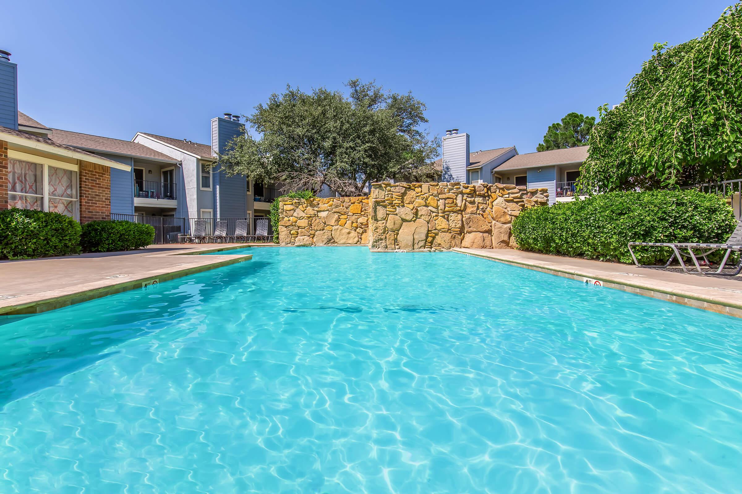 A pristine swimming pool surrounded by stone and greenery, featuring clear blue water under a bright blue sky. Nearby, there are lounge chairs and residential buildings in the background, creating a relaxing outdoor atmosphere.