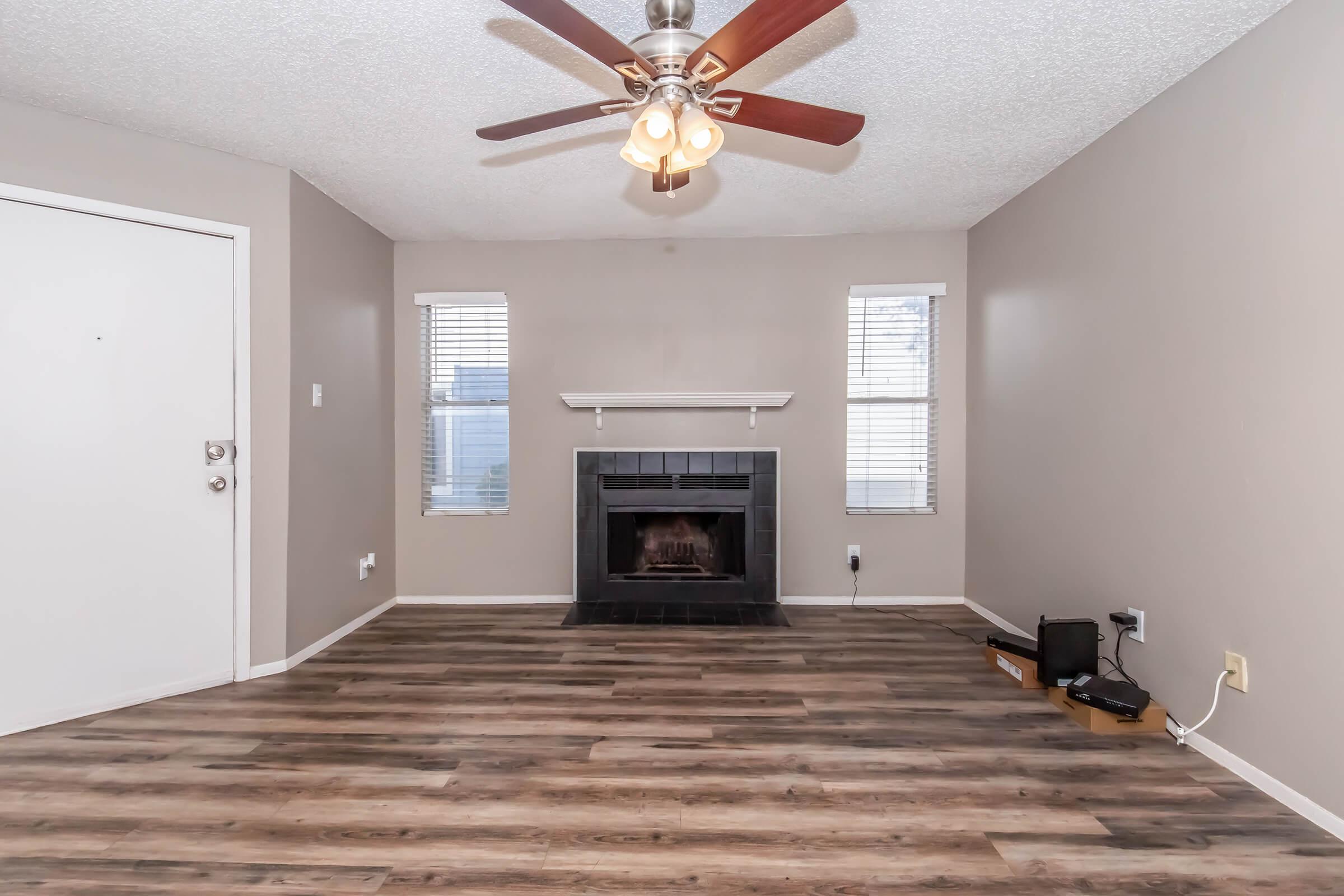 A spacious living room featuring a ceiling fan, a fireplace with a black surround, and large windows letting in natural light. The floor is wooden with a warm tone, and the walls are painted a light beige color. There is a door on the left and some electrical outlets visible.