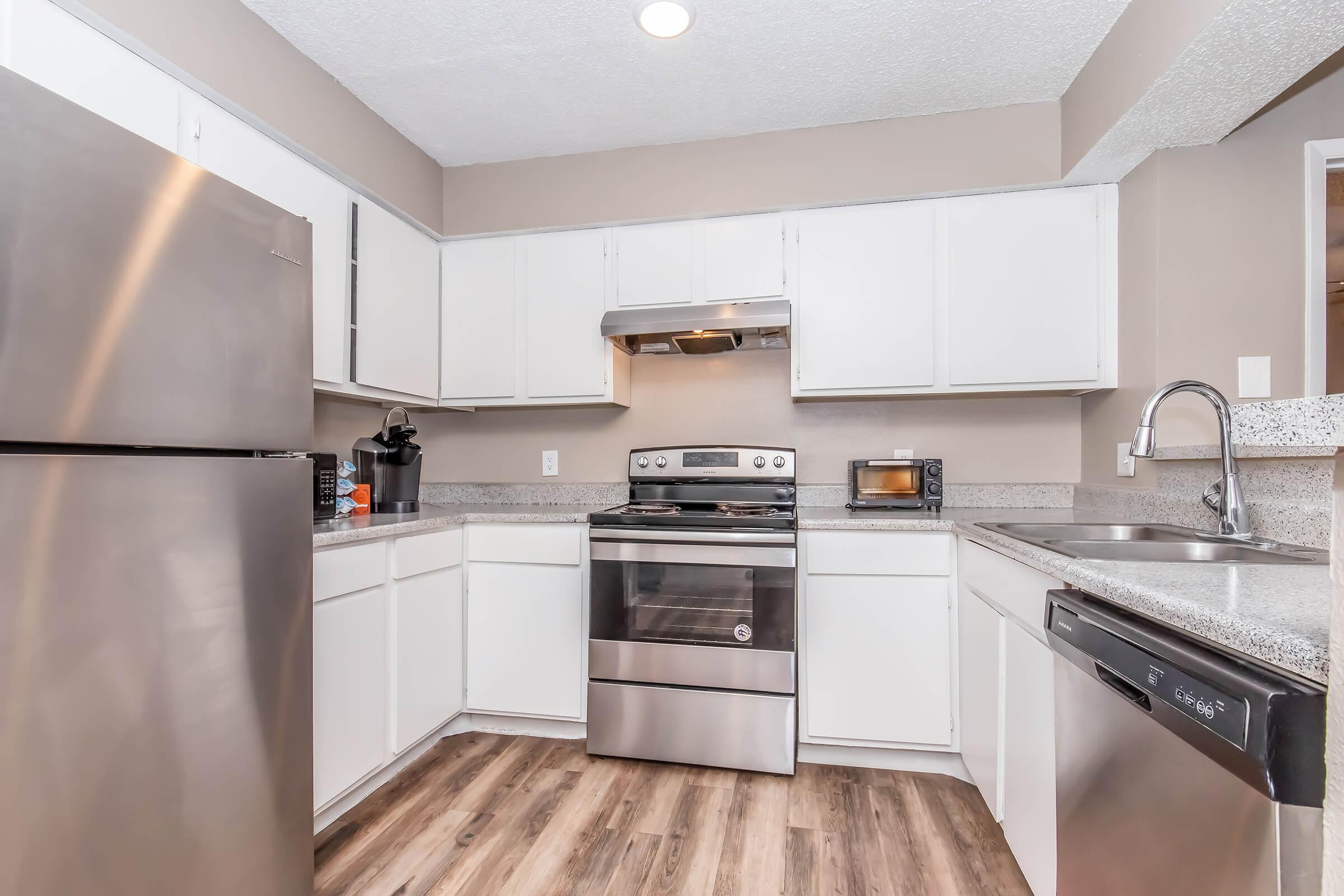 Modern kitchen with stainless steel appliances including a refrigerator, stove, and dishwasher. The countertops are light-colored granite, and there's a built-in microwave. Cabinets are white, and the flooring resembles wood. Natural light enters the space, creating a bright and inviting atmosphere.