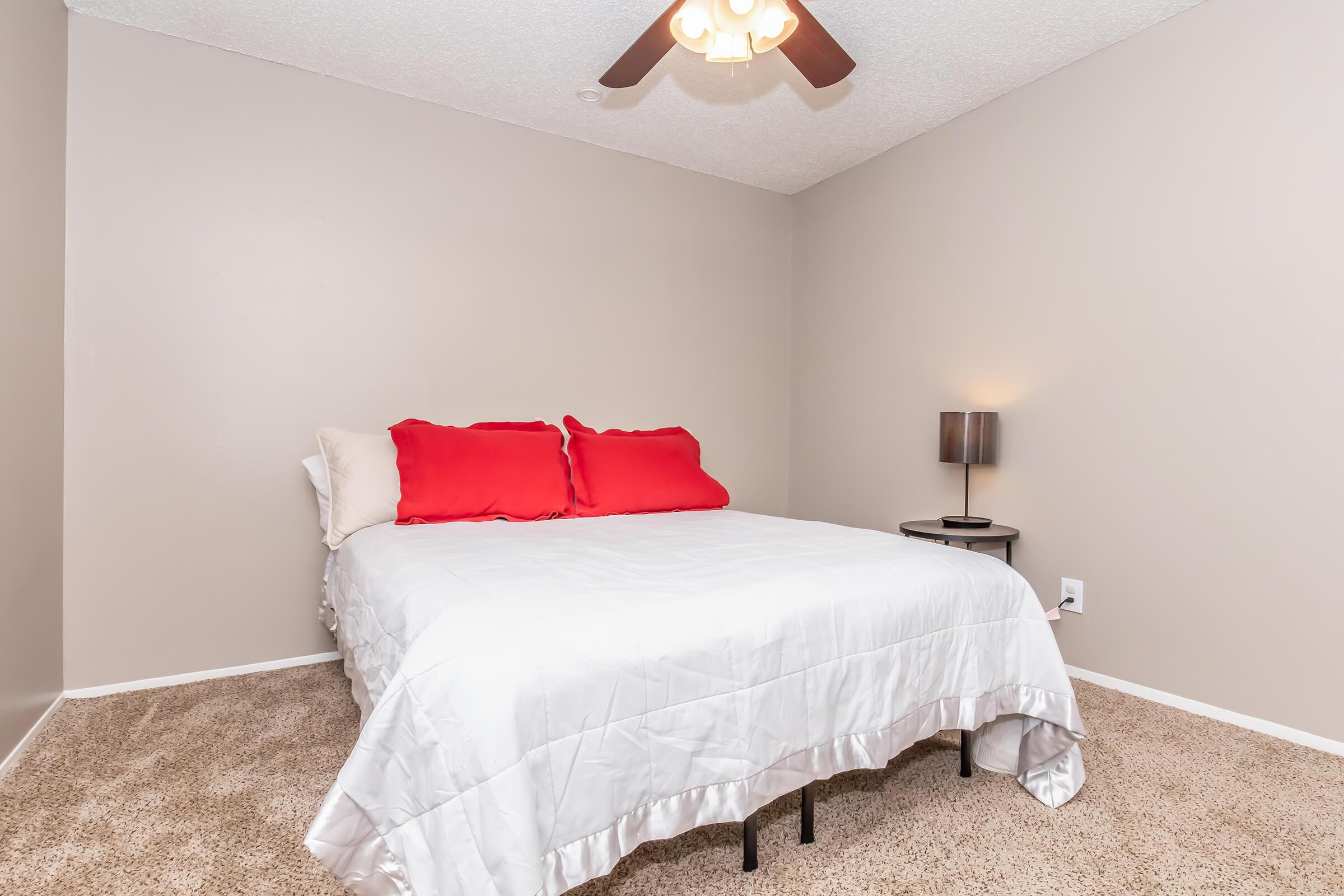 Cozy bedroom featuring a queen-sized bed with a white comforter and vibrant red pillows. There's a small nightstand with a lamp beside the bed. The walls are painted a soft beige, and the room has carpeted flooring. A ceiling fan is visible above, adding to the room's comfort.