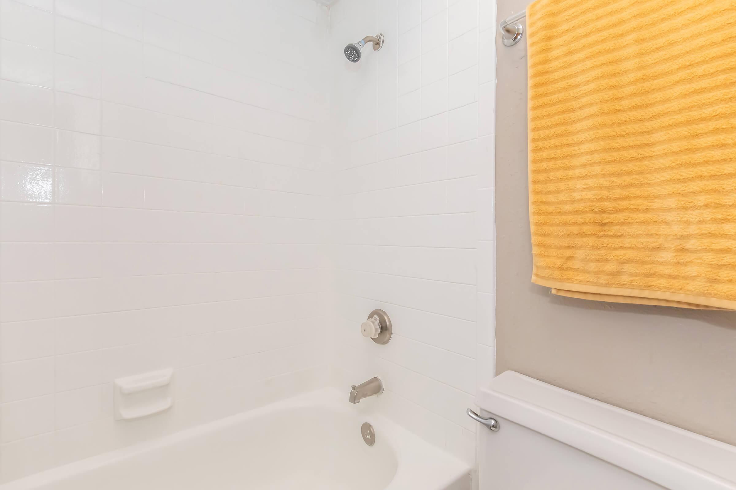 A clean and bright bathroom featuring a white tiled shower area with a chrome showerhead, a white bathtub, and a yellow towel hanging on the wall. The overall decor is minimalistic and modern, focusing on functionality and simplicity.