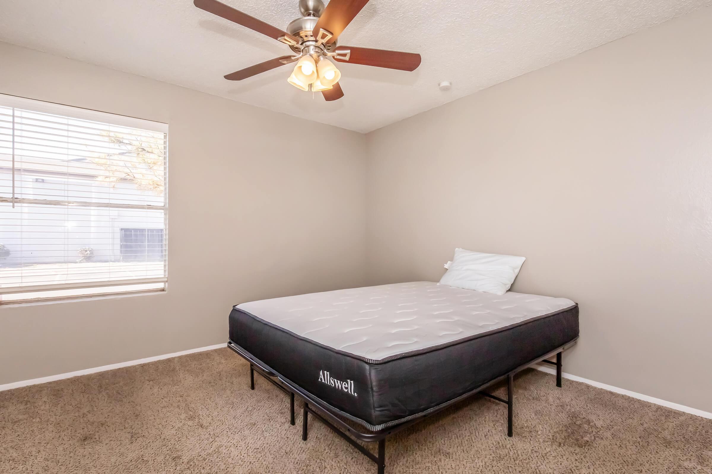 A simple bedroom featuring a queen-sized mattress on a bed frame, a ceiling fan with lights, and a window with blinds. The walls are painted in a neutral color, and the floor is covered with carpet. A pillow is placed on the mattress, adding to the room's minimalistic decor.