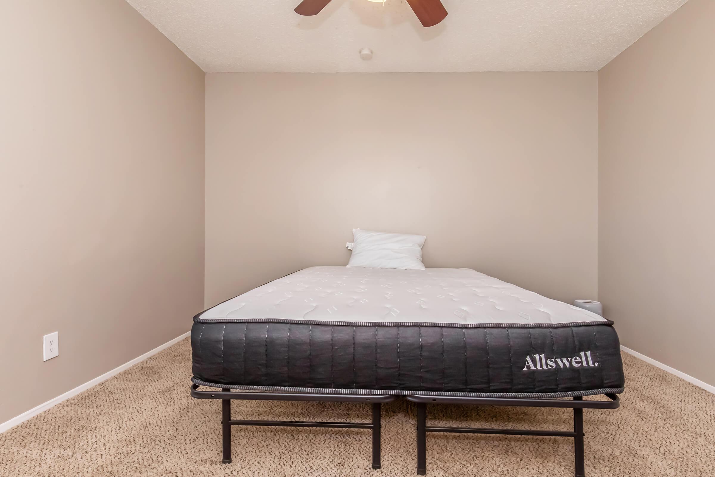 A simple bedroom featuring a single bed with a mattress and a white pillow. The walls are painted in a neutral color, and there is a ceiling fan above. The floor is carpeted in a beige tone, creating a minimalist and clean aesthetic.