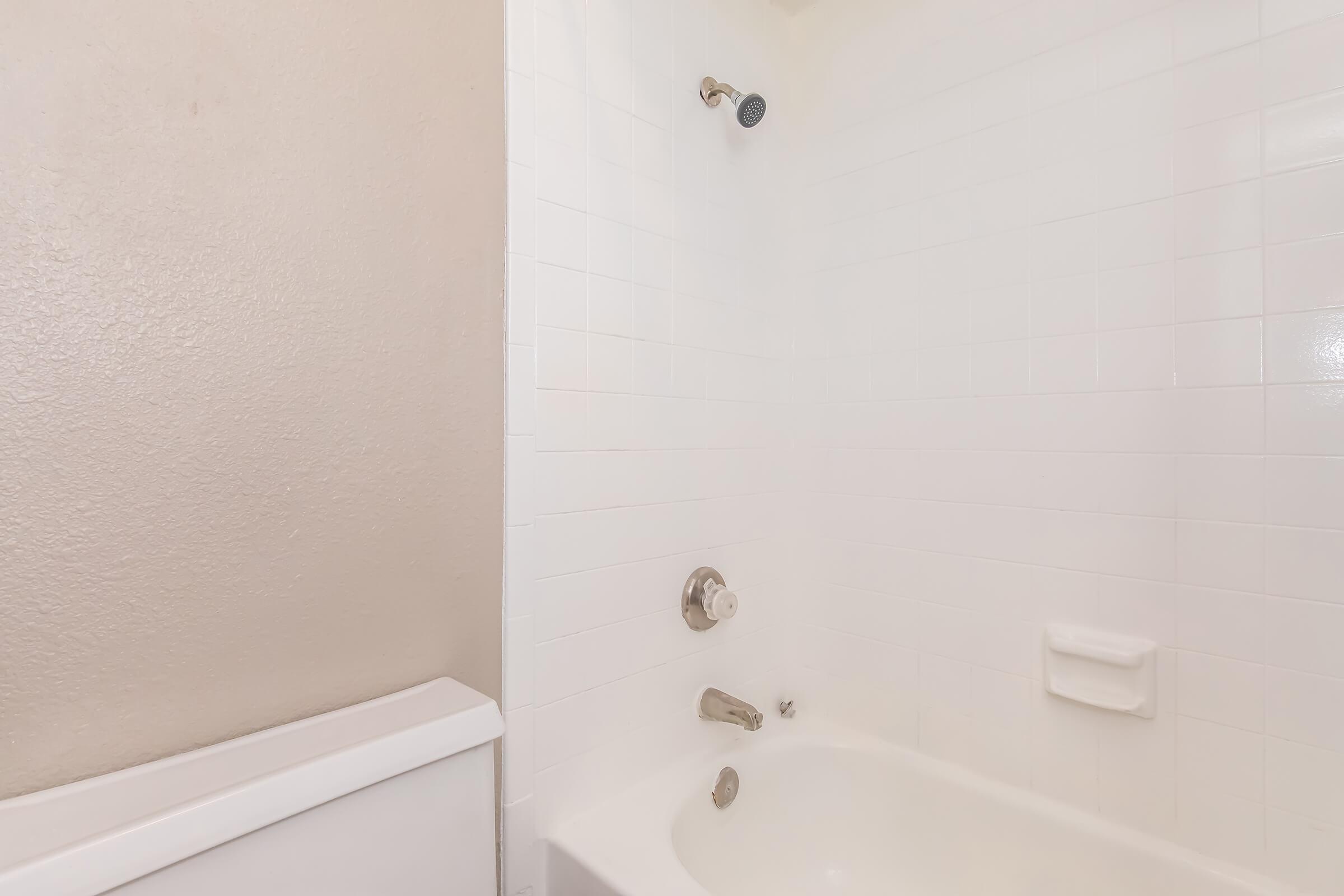 A clean and modern bathroom featuring a white tiled shower and bathtub. The showerhead is mounted on the wall, and there are simple fixtures for hot and cold water controls. The wall adjacent to the bathtub is a neutral color, creating a bright and airy atmosphere.