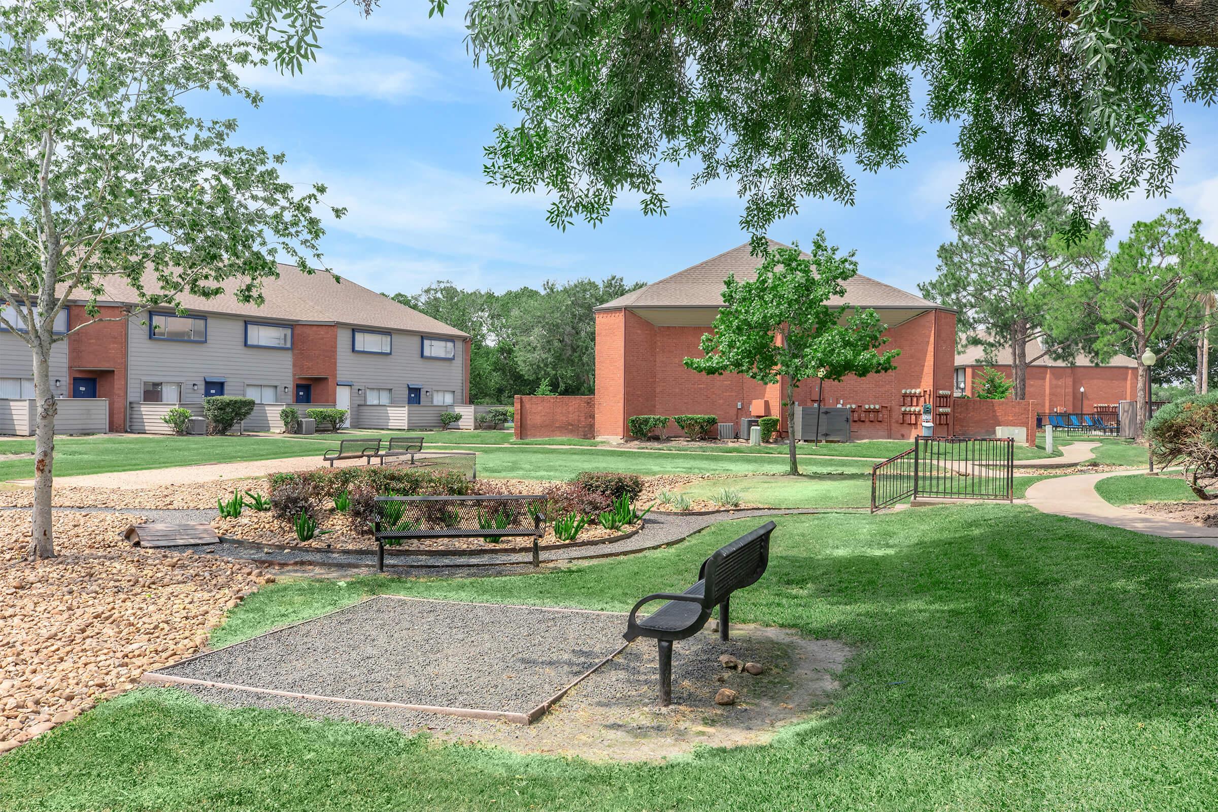 A peaceful outdoor area featuring a grassy landscape with a bench, garden beds, and a small paved path. Surrounding the area are brick buildings and trees, creating a serene environment suitable for relaxation and community gatherings.